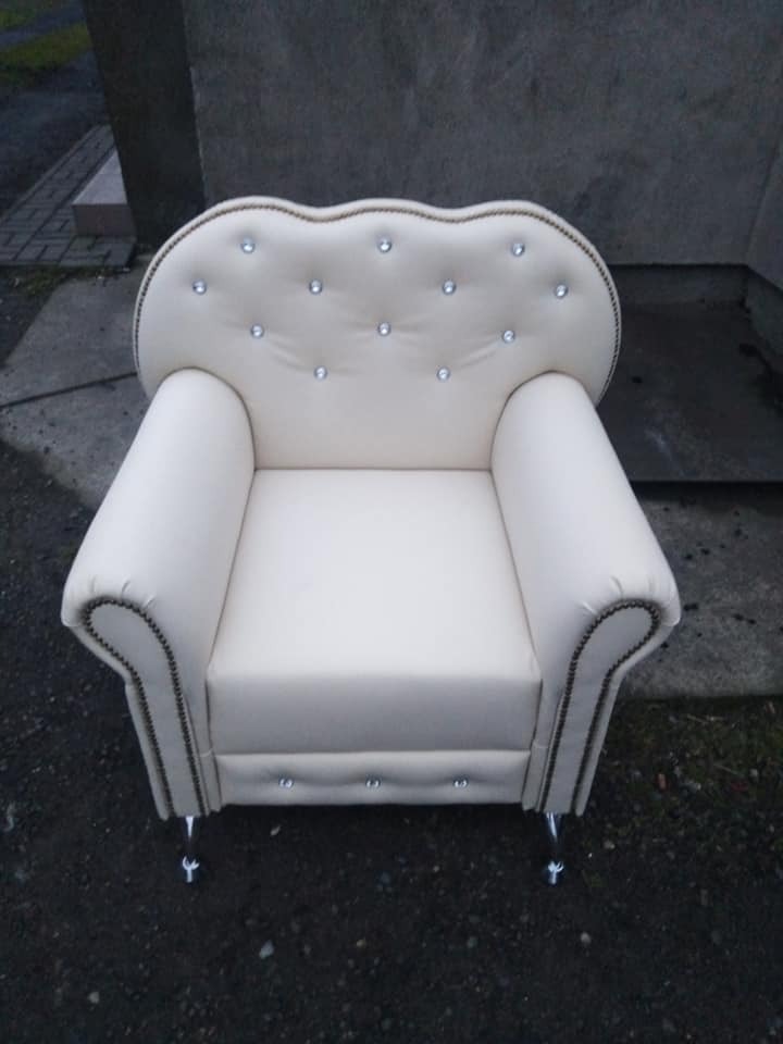 Elegancki, kremowy fotel tapicerowany z ozdobnymi kryształkami i srebrnymi nóżkami, ustawiony na zewnątrz. Widok z przodu, podkreślający detale wykończenia.