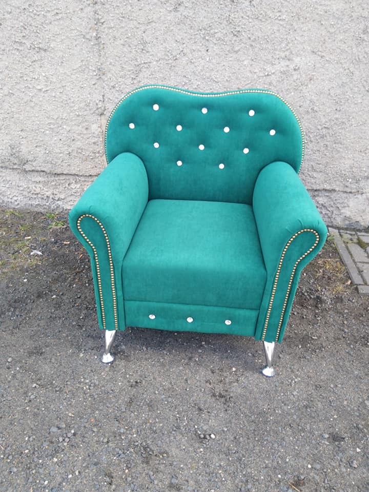 Szmaragdowy fotel z dekoracyjnymi pinezkami i srebrnymi nóżkami na tle szarej ściany. Fotel tapicerowany, z guzikami na oparciu i pod siedziskiem.