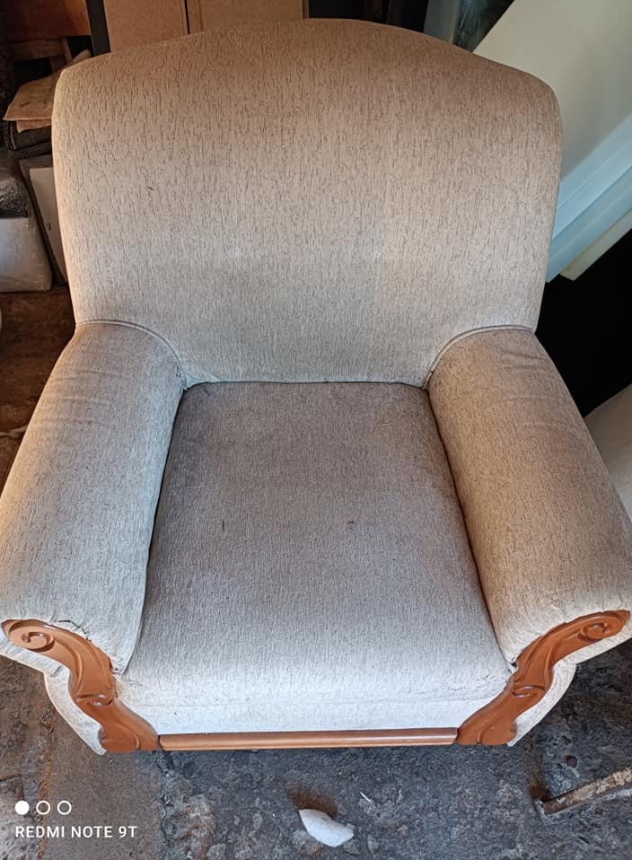 Beżowy fotel tapicerowany z drewnianymi zdobieniami na podłokietnikach, sfotografowany z przodu, ukazujący detale tkaniny i rzeźbione elementy.