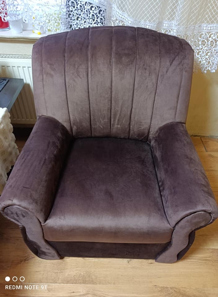 Brązowy, welurowy fotel z wysokim oparciem i zaokrąglonymi podłokietnikami, stojący na drewnianej podłodze. Widoczne detale tapicerki i dekoracyjna zasłona w tle.