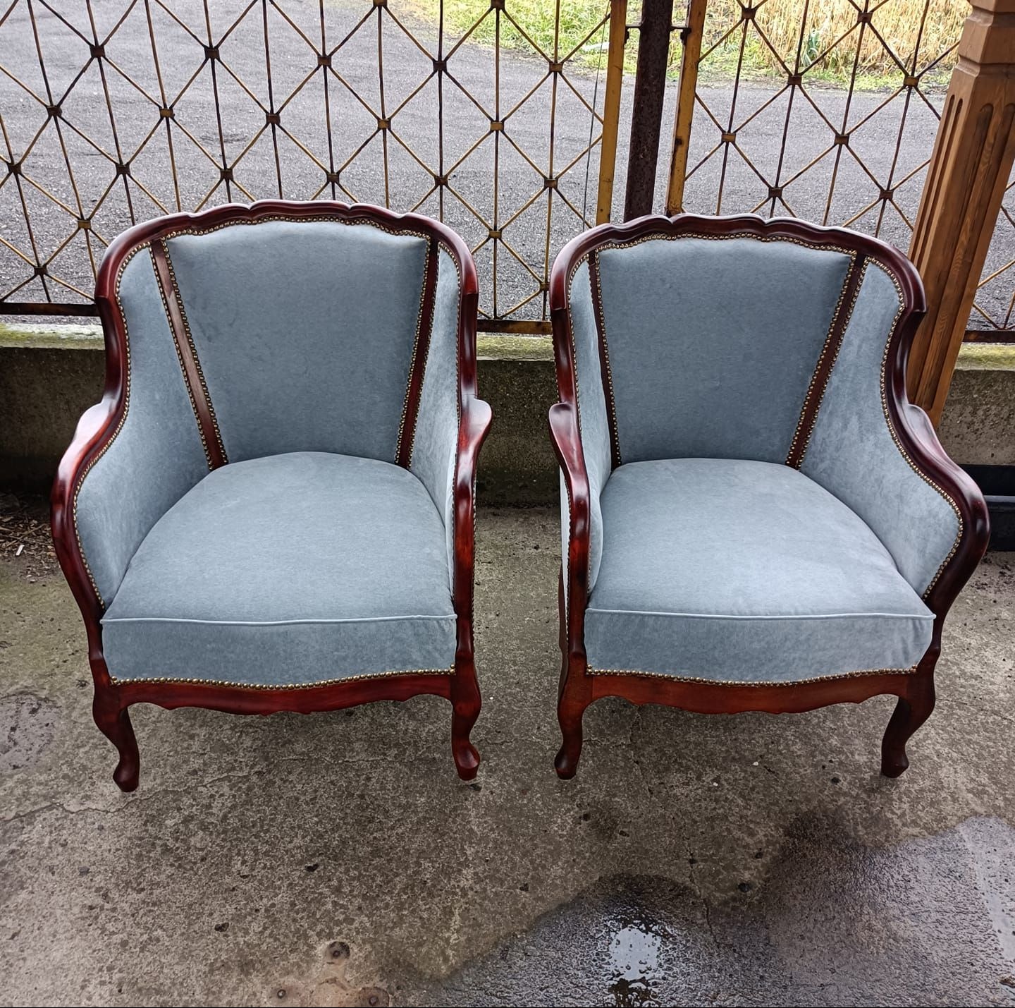 Dwa odnowione fotele w stylu vintage z drewnianymi ramami i szarą tapicerką, ustawione na zewnątrz na betonowym podłożu, z metalowym ogrodzeniem w tle.