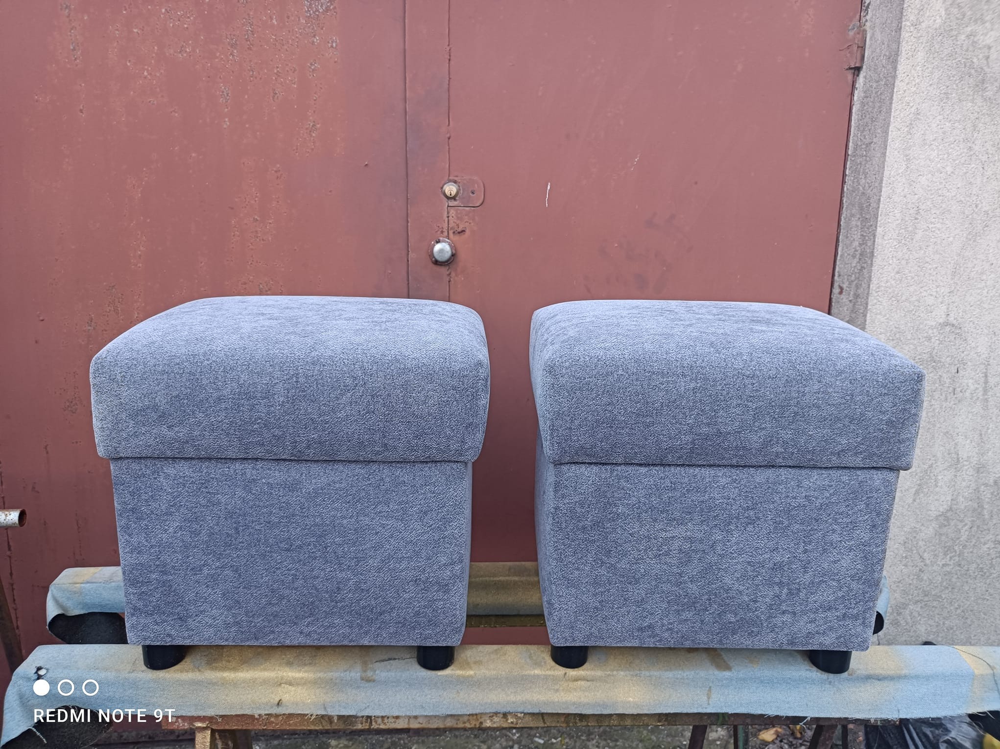 Dwa identyczne, szare, tapicerowane pufy na niskich nóżkach, ustawione obok siebie na tle czerwonych, metalowych drzwi garażowych. Widoczne detale tapicerki i konstrukcji.