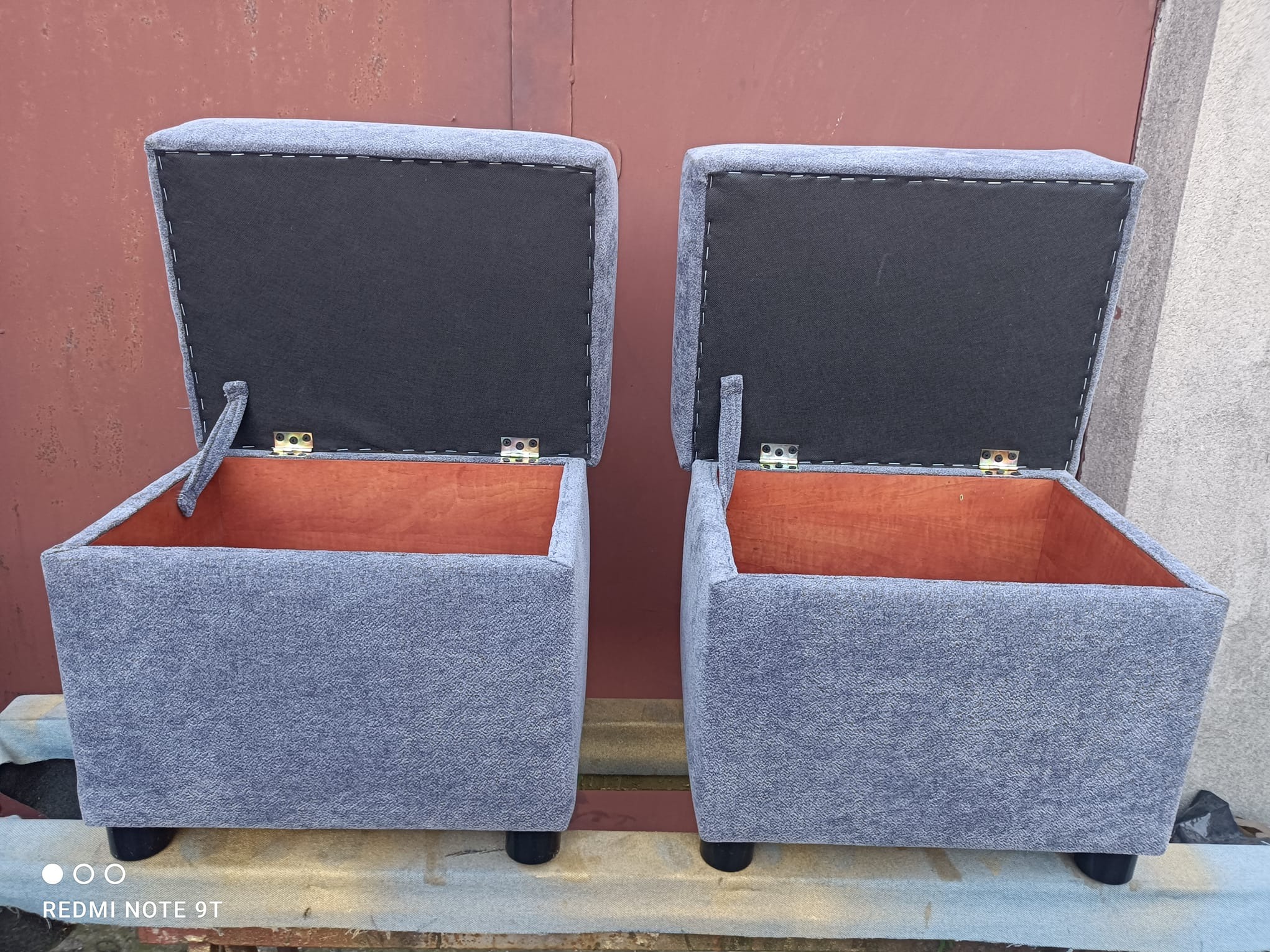 Dwa otwarte, szare pufy tapicerowane z drewnianym wnętrzem, ustawione obok siebie na zewnątrz. Widoczne zawiasy i czarne nóżki. Tło stanowi blaszana ściana.