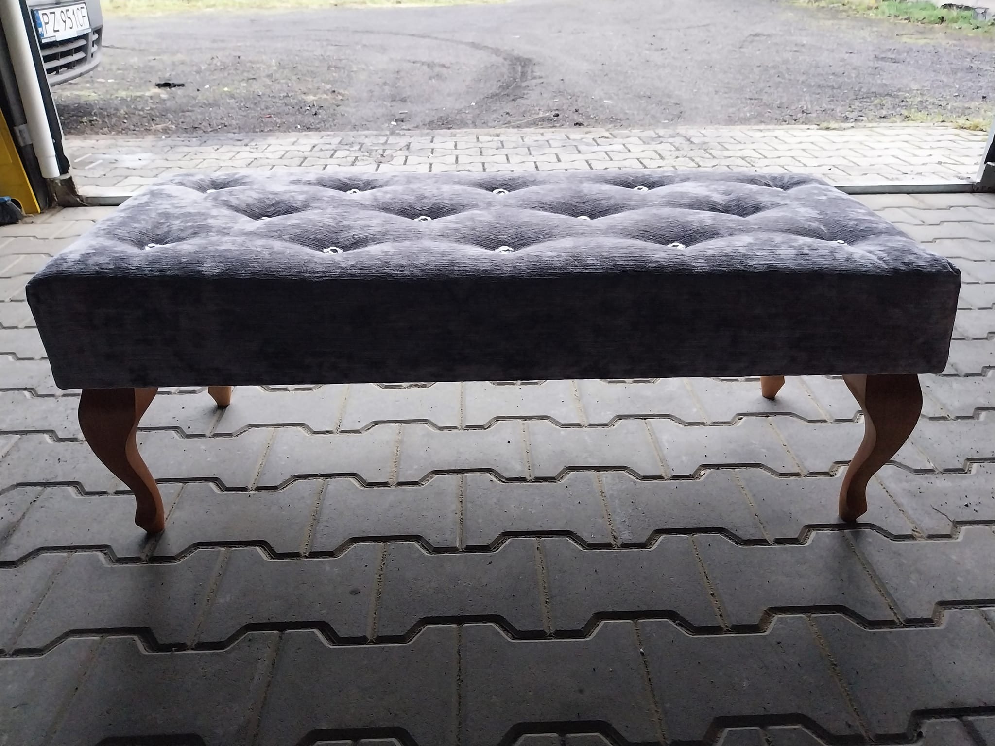 Szary, tapicerowany puf z guzikami i drewnianymi nóżkami, ustawiony na szarej kostce brukowej. Widok z przodu, oświetlenie naturalne.