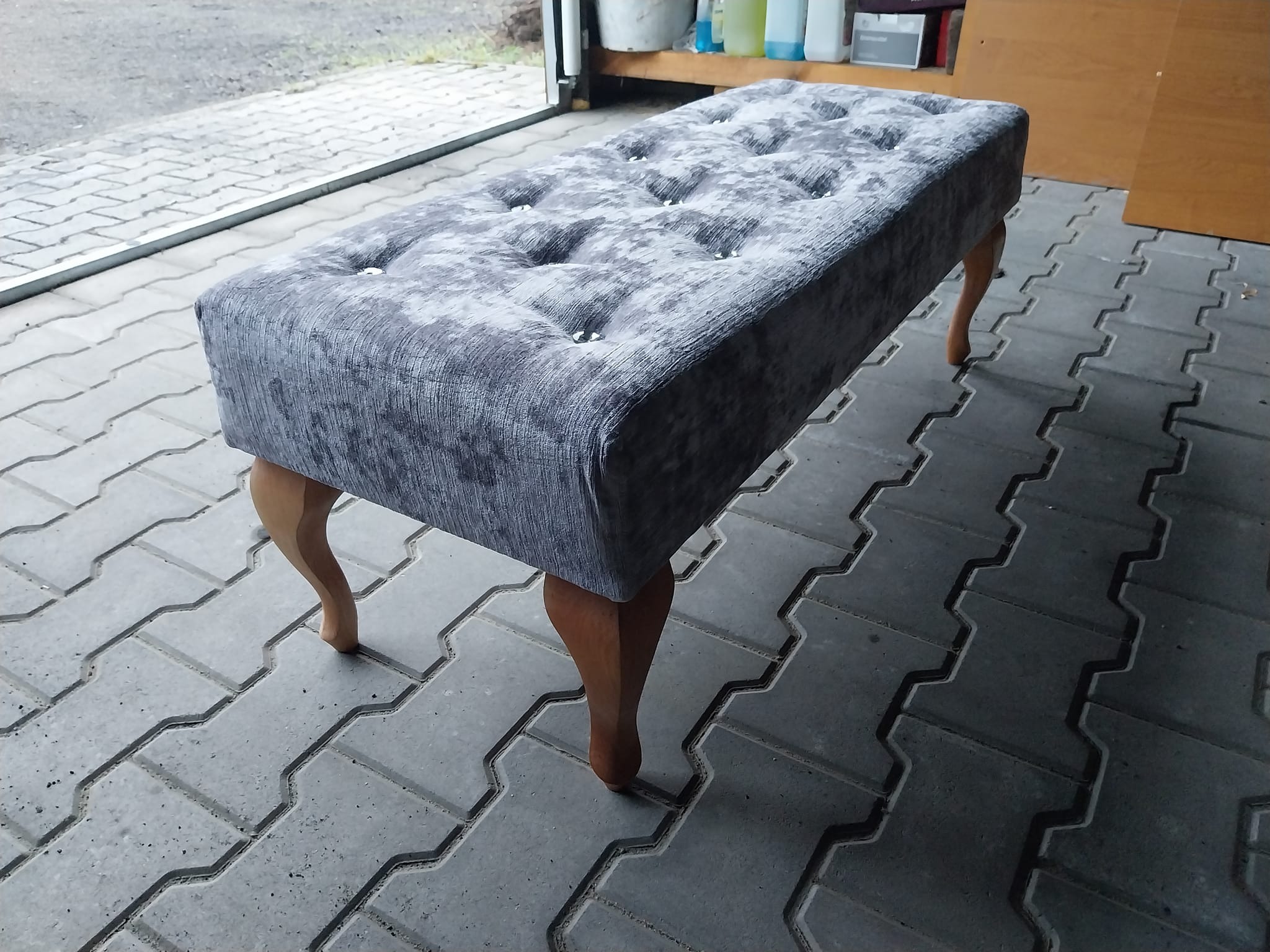 Tapicerowana ławka w odcieniach szarości z drewnianymi nóżkami, pikowana z guzikami, ustawiona na szarej kostce brukowej, w tle widoczne przedmioty.