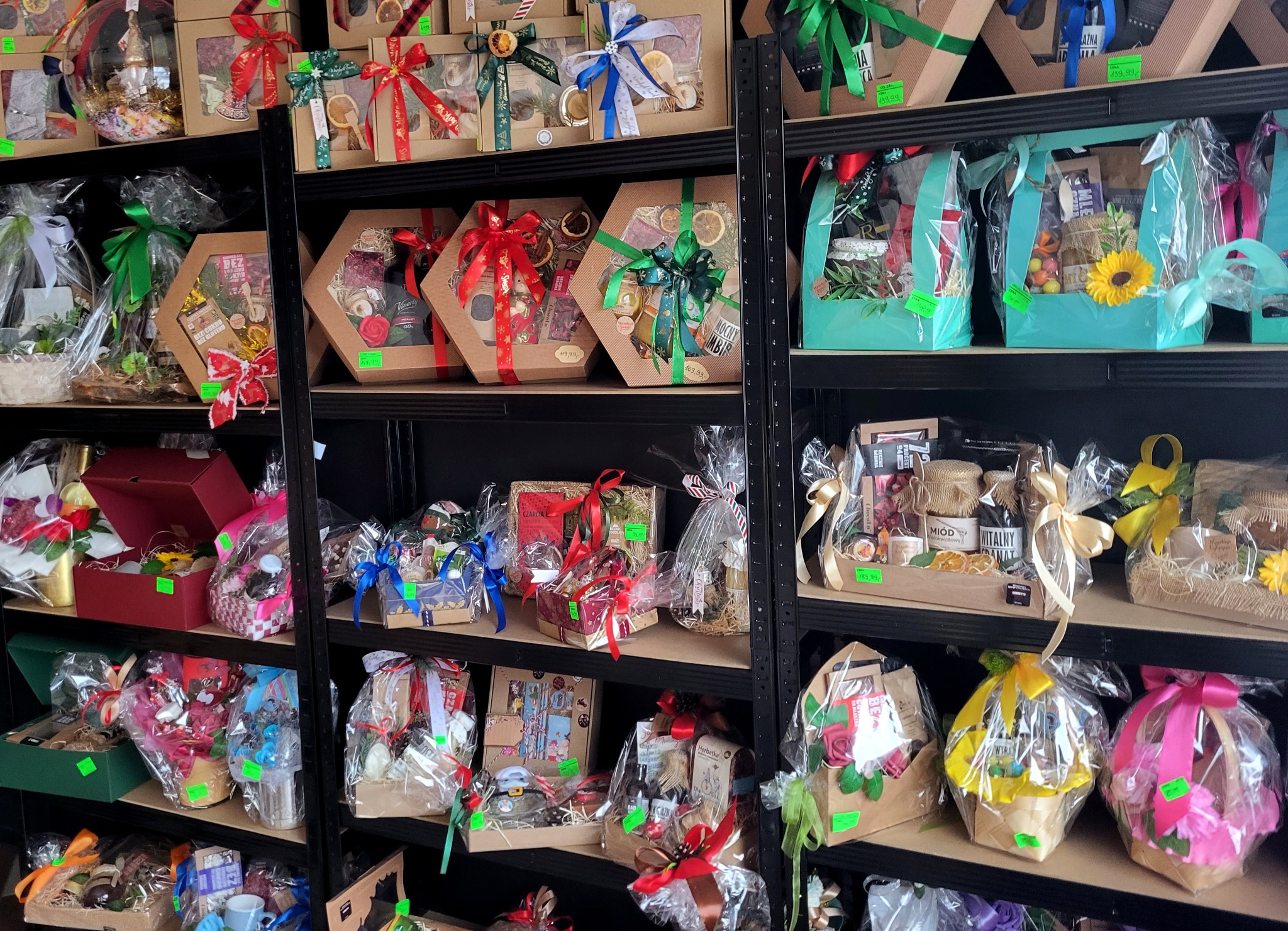 Regał z różnorodnymi koszami prezentowymi, zapakowanymi w celofan i ozdobionymi wstążkami. Widoczne pudełka sześciokątne i kosze z zawartością, np. miód, słodycze, herbata.