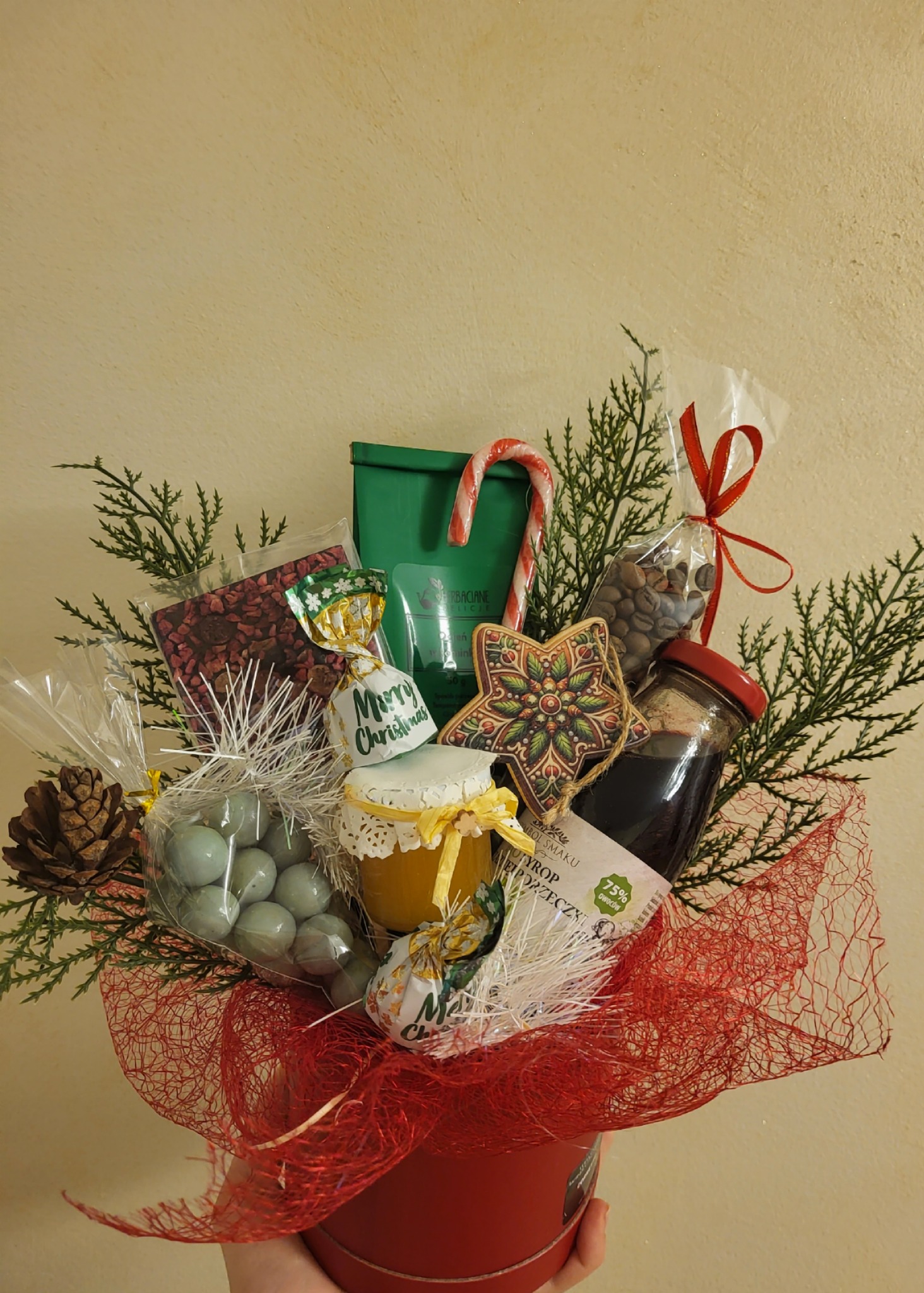 Ręcznie robiony kosz prezentowy w czerwonym pudełku z dekoracjami świątecznymi, słodyczami, miodem i kawą, owinięty czerwoną siatką i gałązkami świerku.