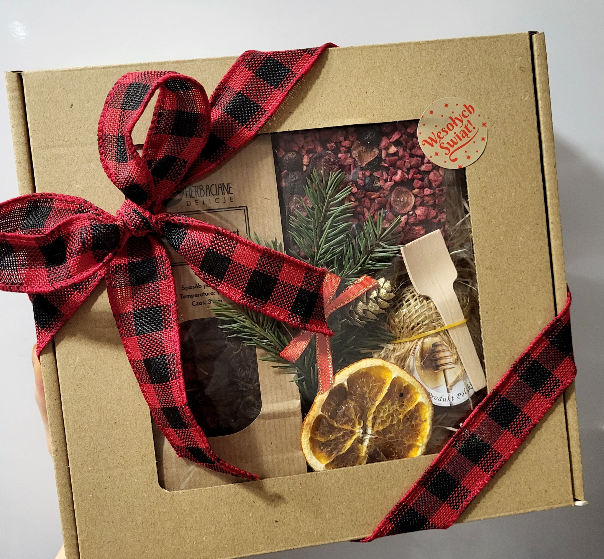 Kartonowy kosz prezentowy z okienkiem, wypełniony herbatą, miodem i suszonymi owocami, ozdobiony czerwoną wstążką w kratę i naklejką 'Wesołych Świąt!'.