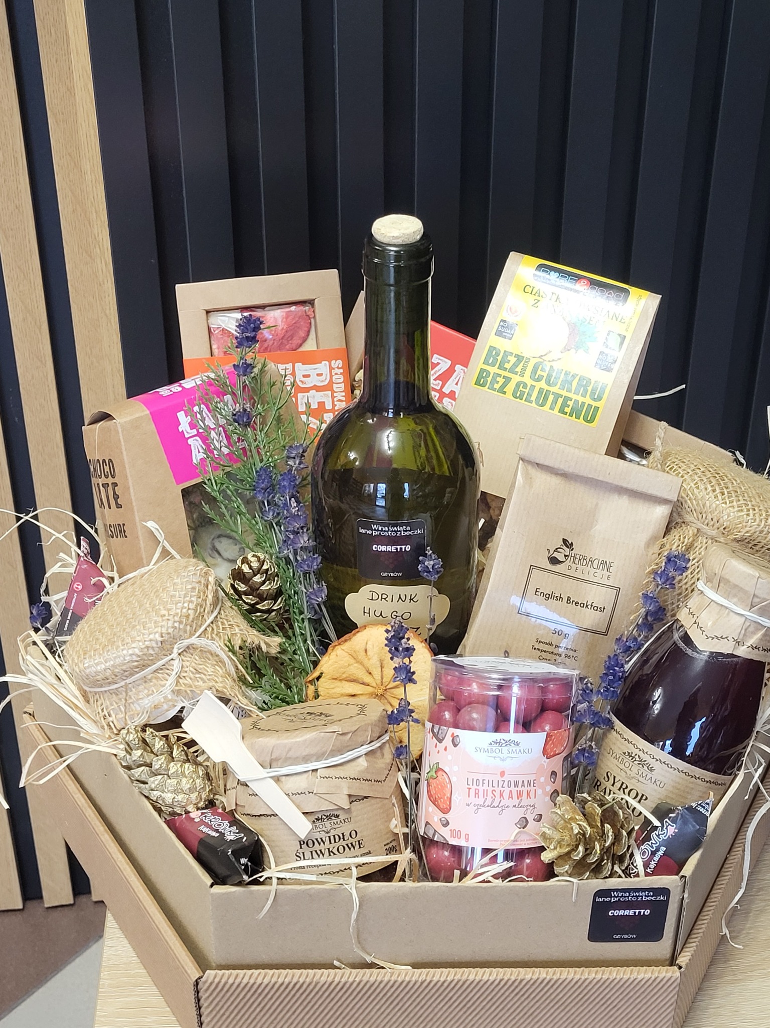 Elegancki kosz prezentowy z winem, słodyczami, herbatą i powidłami, udekorowany lawendą i szyszkami, na tle ciemnej ściany z drewnianymi panelami.