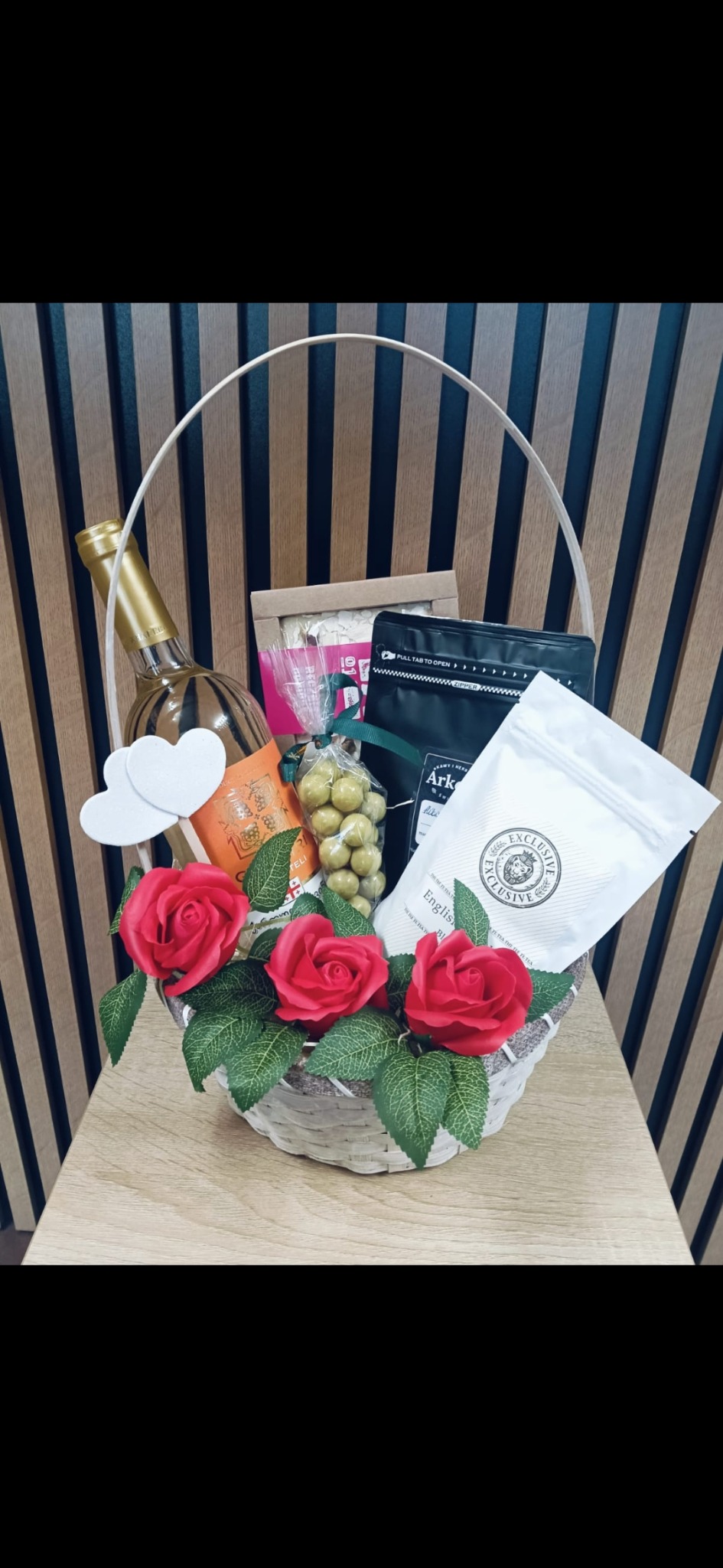 Elegancki kosz prezentowy z winem, słodyczami i dekoracjami kwiatowymi na drewnianym stole, na tle ściany z pionowych paneli. Idealny upominek na specjalną okazję.