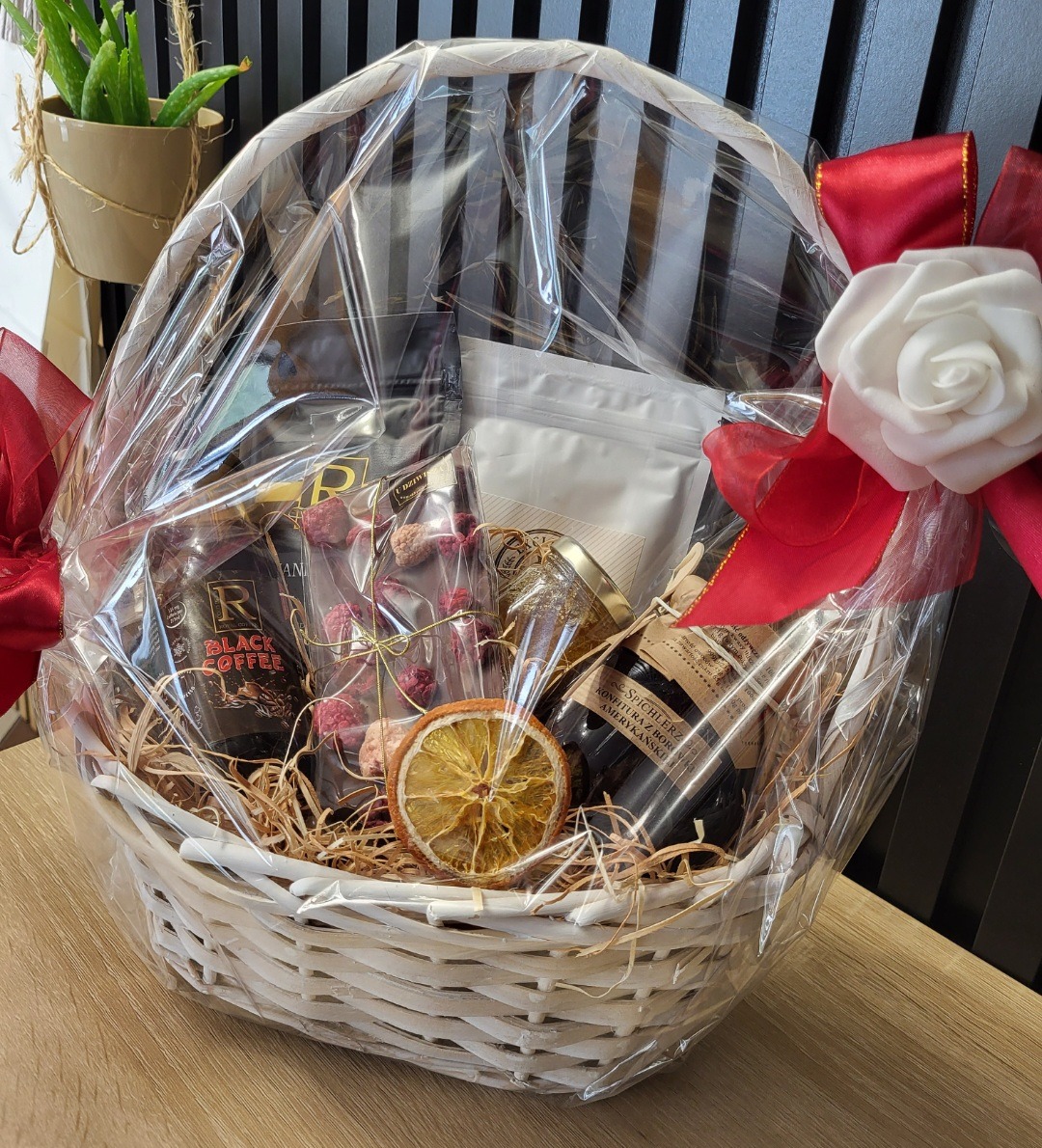 Elegancki kosz prezentowy z kawą, czekoladą, konfiturą i suszoną pomarańczą, owinięty folią i ozdobiony czerwoną wstążką z białą różą.