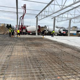 PATRON-STAL JAKUB PARCEJ - Zbrojenie pod wylewkę fundamentów hali przemysłowej. Widoczne betoniarki i pracownicy w odblaskowych kamizelkach. Konstrukcja stalowa dachu w tle.