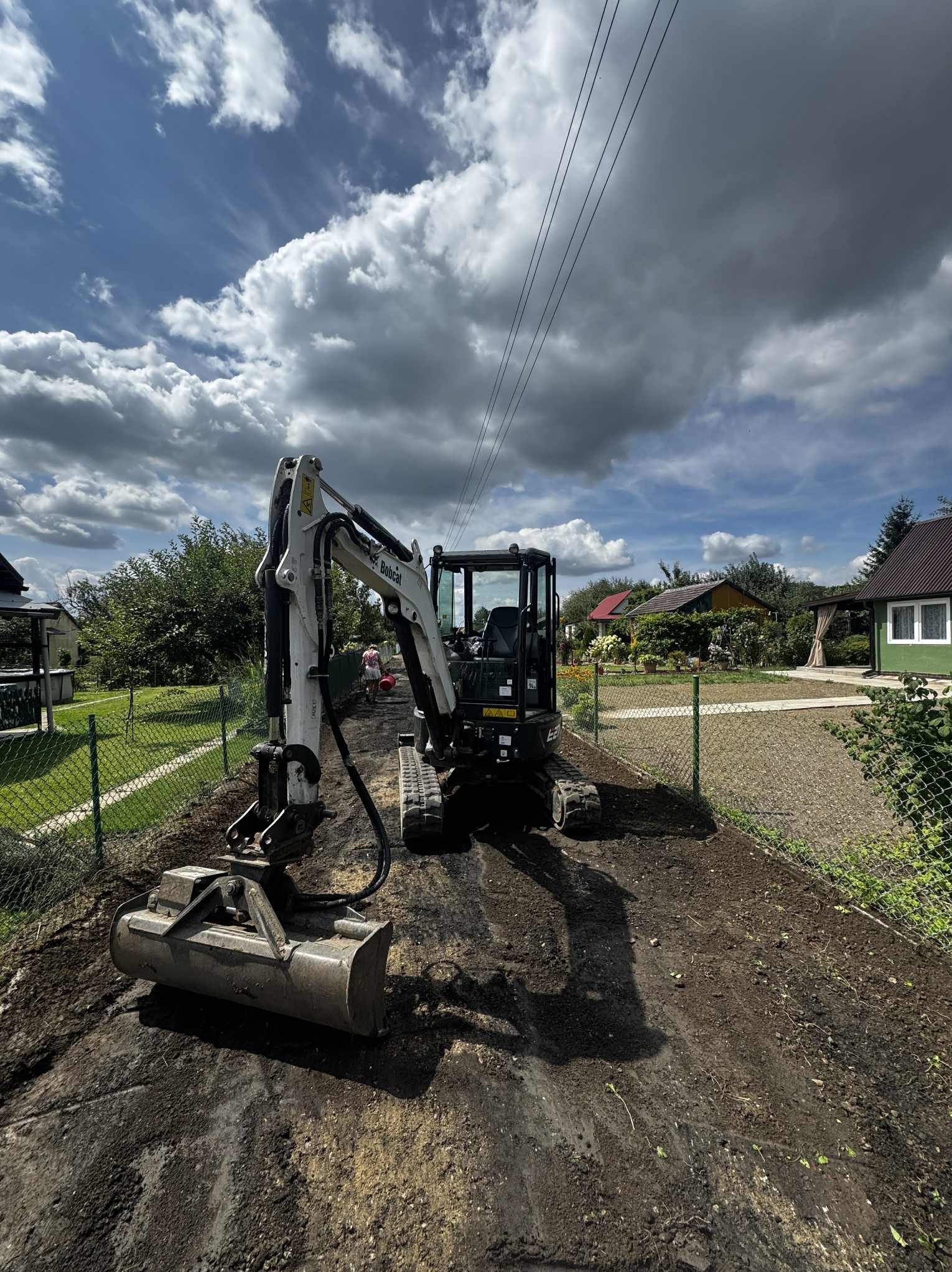 Mini koparka Bobcat na działce, przygotowuje teren pod budowę. Widoczne ogrodzenie z siatki i domy w tle. Pochmurne niebo.