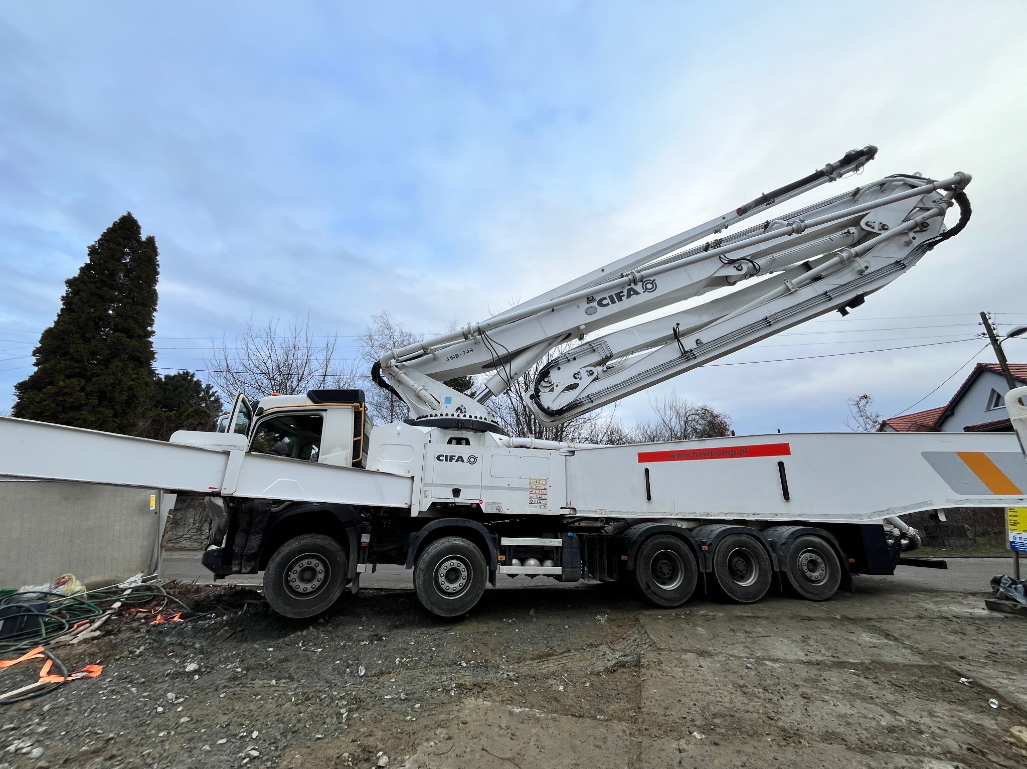 Betonomieszarka CIFA na placu budowy, z wysięgnikiem skierowanym ku górze, obok widoczny fragment betonowego muru i nieporządek budowlany.