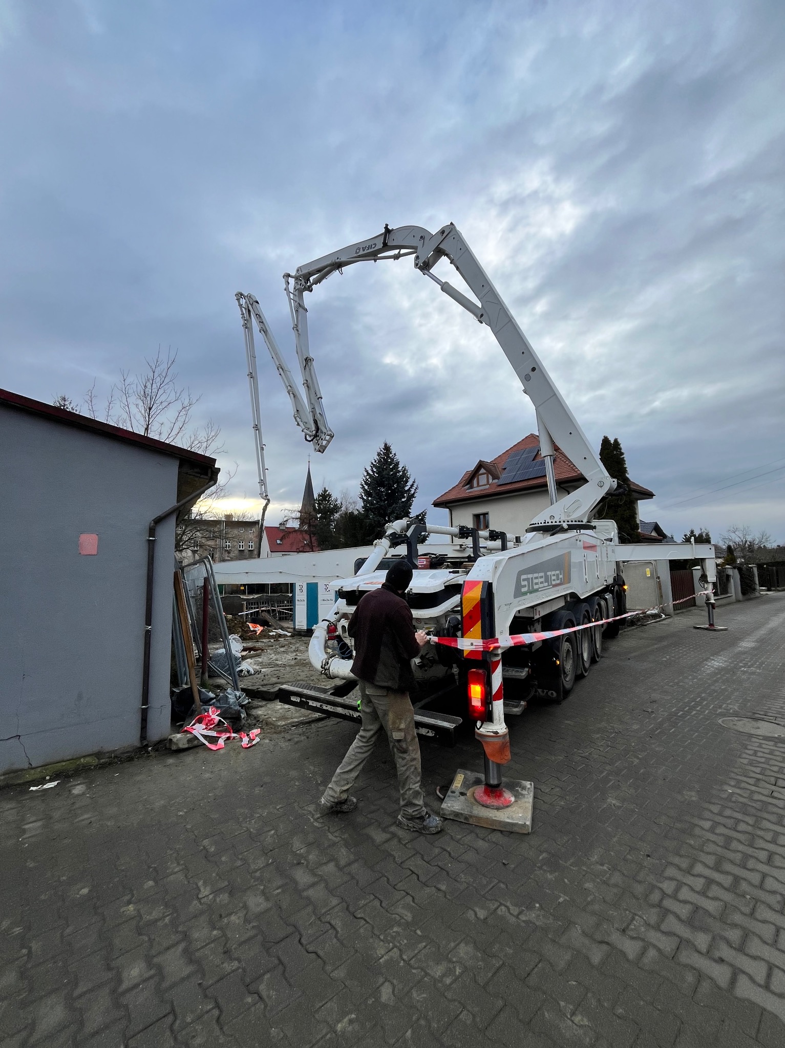 Betonomieszarka Steeltech na budowie, z wysięgnikiem nad terenem. Pracownik w roboczym ubraniu nadzoruje proces. W tle budynki i pochmurne niebo.