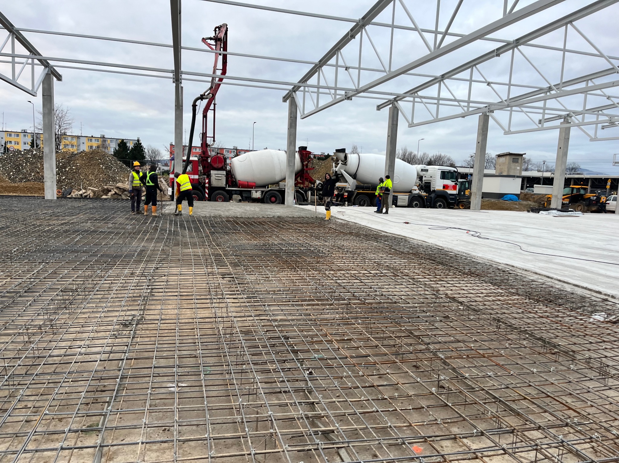 Zbrojenie pod wylewkę fundamentów hali przemysłowej. Widoczne betoniarki i pracownicy w odblaskowych kamizelkach. Konstrukcja stalowa dachu w tle.