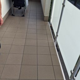 REM-BUD ROBERT GŁOGOWSKI - Wyłożony brązowymi płytkami balkon z białą balustradą i suszarką na ubrania. Widok z góry, ukazujący regularny wzór płytek i minimalistyczny design balustrady.