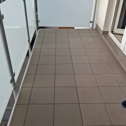 REM-BUD ROBERT GŁOGOWSKI - Kafelki na balkonie z białymi szklanymi barierkami i metalowymi słupkami. Widok z góry na brązowe płytki podłogowe. Fragment buta w prawym dolnym rogu.