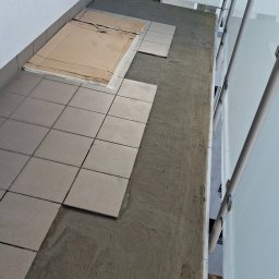 REM-BUD ROBERT GŁOGOWSKI - Układanie szarych płytek na balkonie, widoczne fragmenty kartonu ochronnego i świeża zaprawa. Ujęcie z góry, z widoczną balustradą.