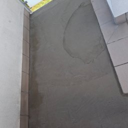 REM-BUD ROBERT GŁOGOWSKI - Wyrównana, szara wylewka na balkonie, przygotowana pod dalsze prace wykończeniowe. Widoczne płytki na krawędzi i fragment zieleni w tle.