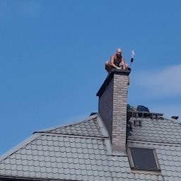 REM-BUD ROBERT GŁOGOWSKI - Pracownik w kucki na szczycie ceglanego komina, na tle błękitnego nieba. Widoczny fragment dachu z szarej dachówki i okno dachowe.