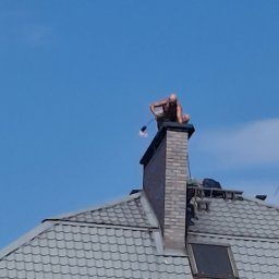 REM-BUD ROBERT GŁOGOWSKI - Mężczyzna siedzi na kominie z narzędziem na dachu pokrytym szarą dachówką, na tle błękitnego nieba. Nietypowe ujęcie z dołu.