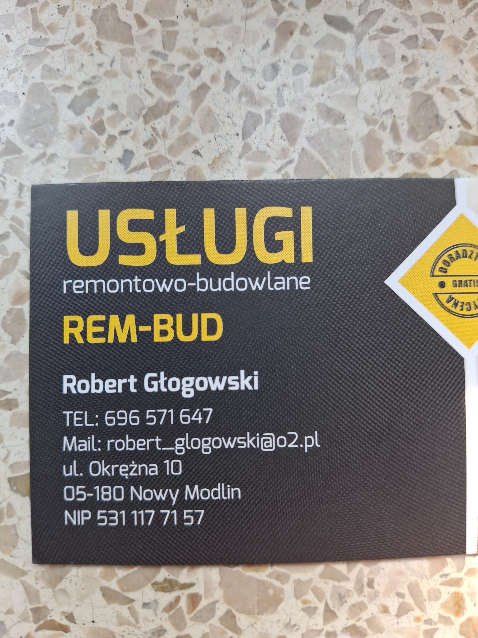 Wizytówka firmy REM-BUD Roberta Głogowskiego oferującej usługi remontowo-budowlane z numerem telefonu, adresem e-mail i NIP-em, leżąca na tle marmurowej posadzki.