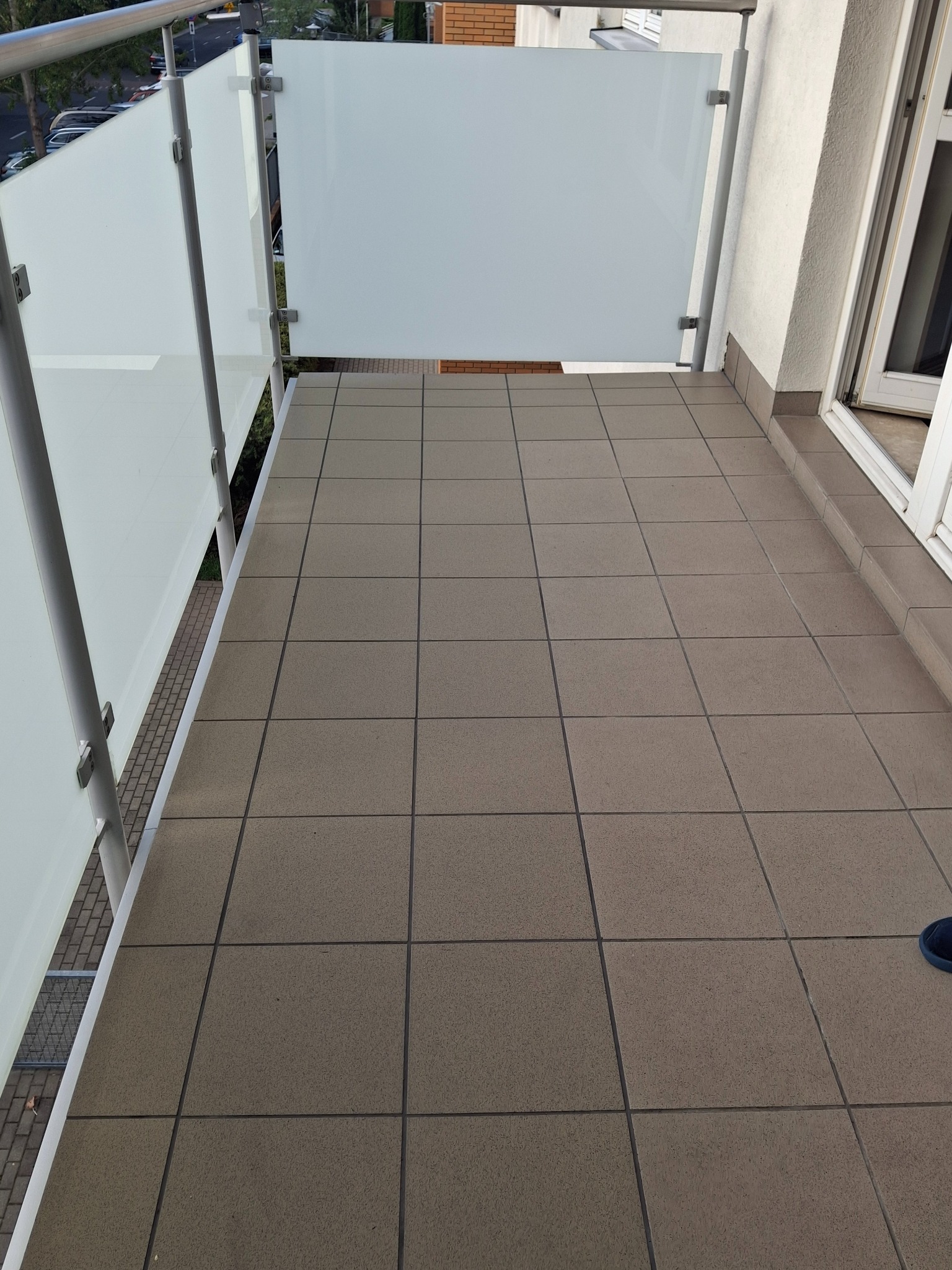 Kafelki na balkonie z białymi szklanymi barierkami i metalowymi słupkami. Widok z góry na brązowe płytki podłogowe. Fragment buta w prawym dolnym rogu.