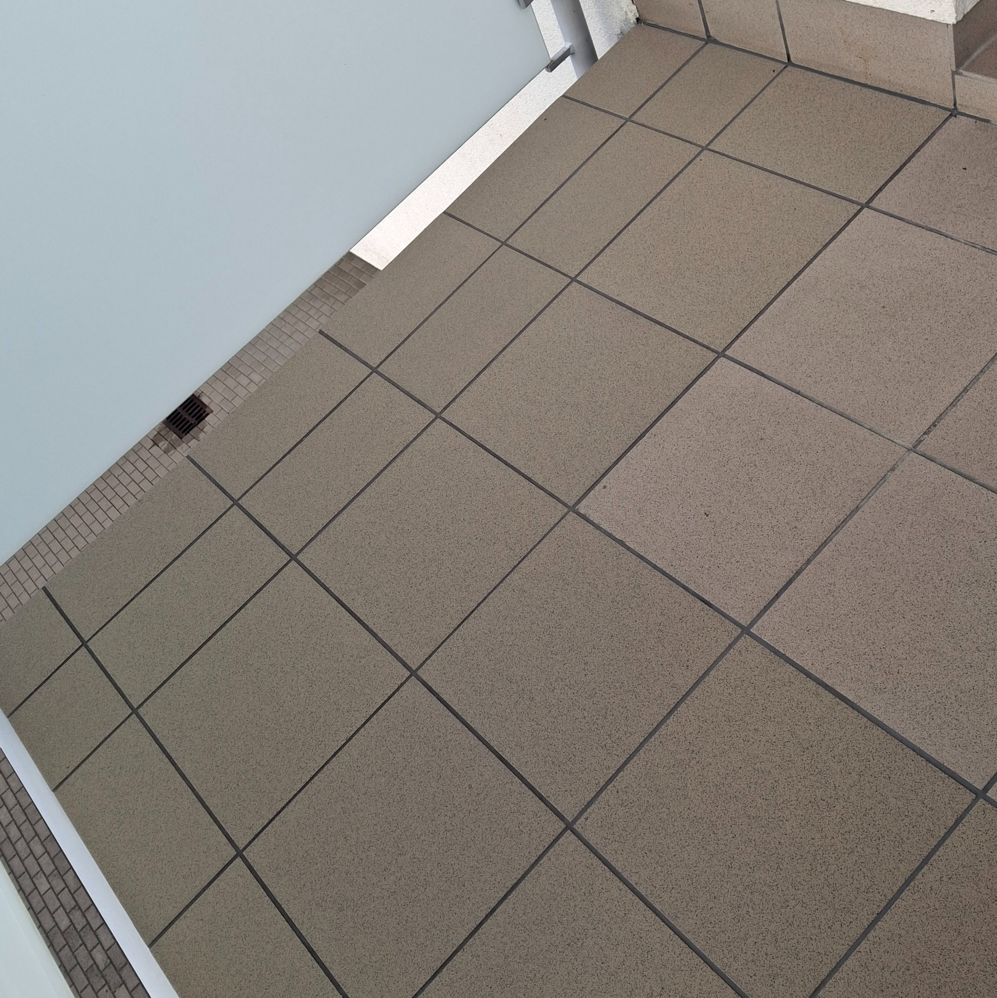 Beżowe płytki podłogowe ułożone na balkonie, widoczne pod kątem. W tle biała ściana i odpływ liniowy. Minimalistyczny design.