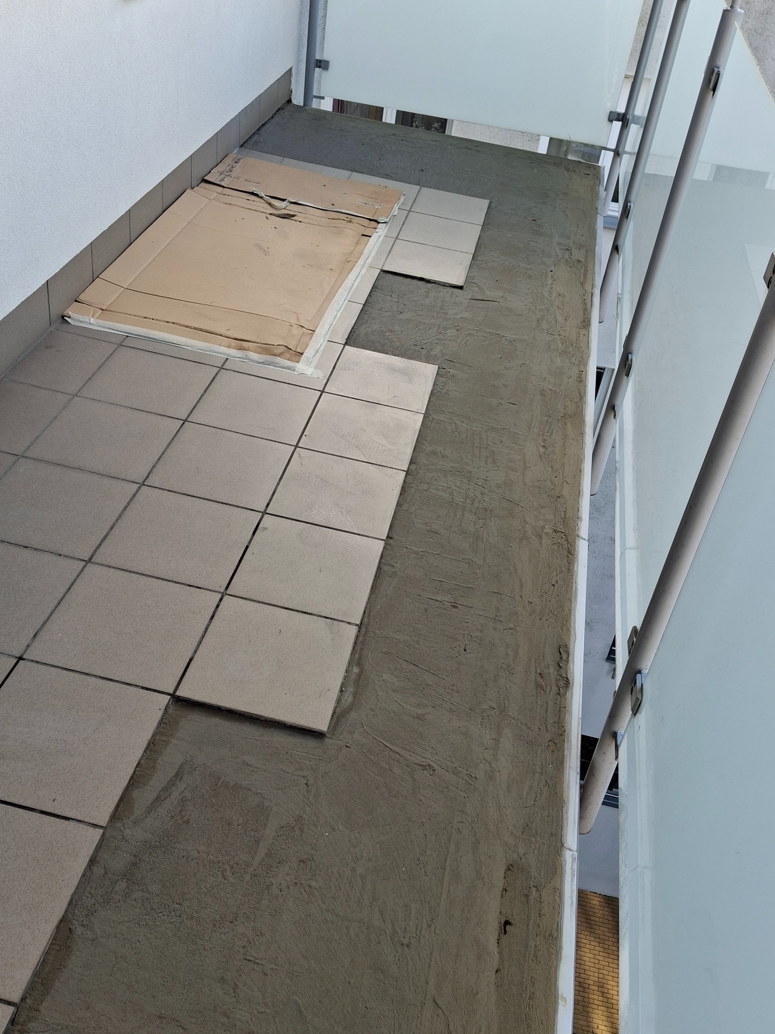 Układanie szarych płytek na balkonie, widoczne fragmenty kartonu ochronnego i świeża zaprawa. Ujęcie z góry, z widoczną balustradą.