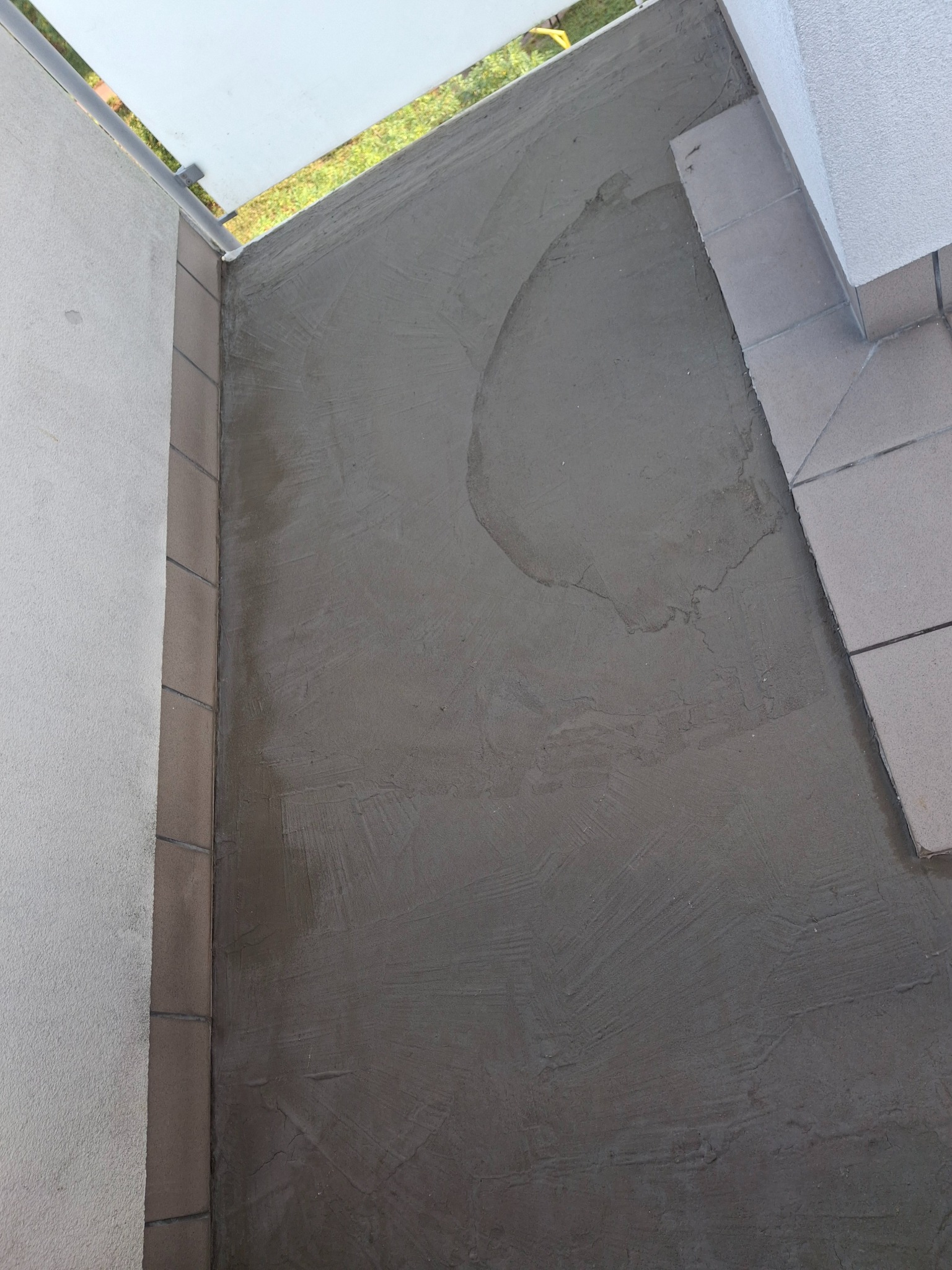 Wyrównana, szara wylewka na balkonie, przygotowana pod dalsze prace wykończeniowe. Widoczne płytki na krawędzi i fragment zieleni w tle.