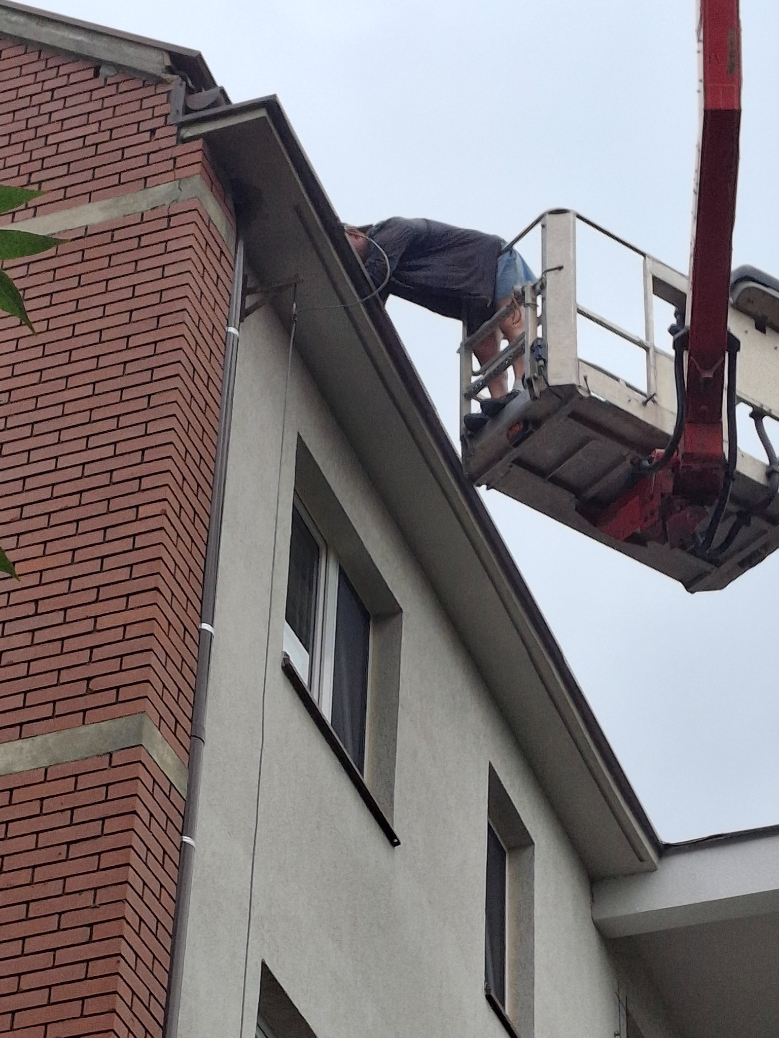 Pracownik na podnośniku koszowym naprawia rynnę przy budynku z ceglaną elewacją i szarym tynkiem. Widoczne okna i fragment zieleni. Ujęcie z dołu.