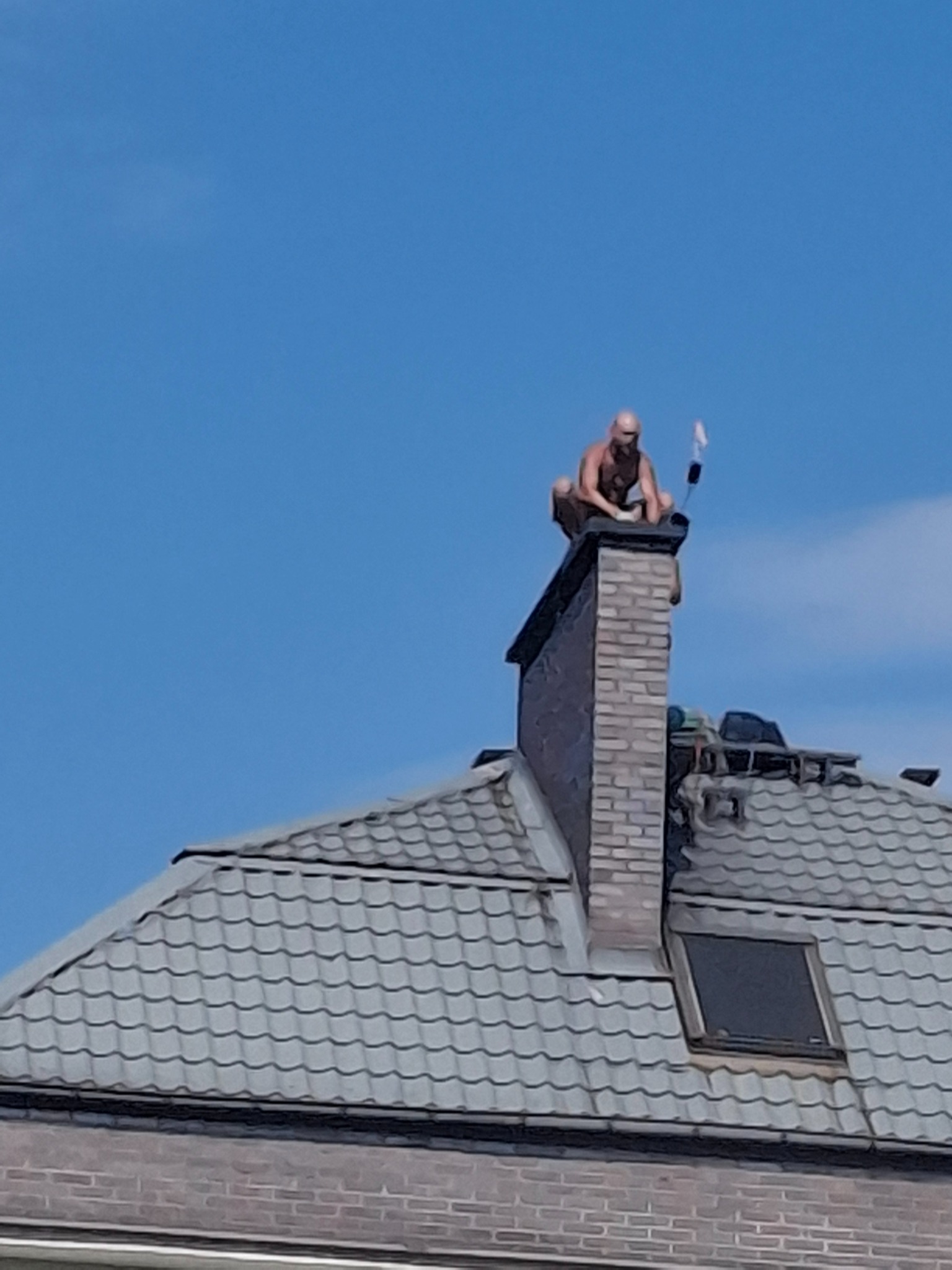 Pracownik w kucki na szczycie ceglanego komina, na tle błękitnego nieba. Widoczny fragment dachu z szarej dachówki i okno dachowe.
