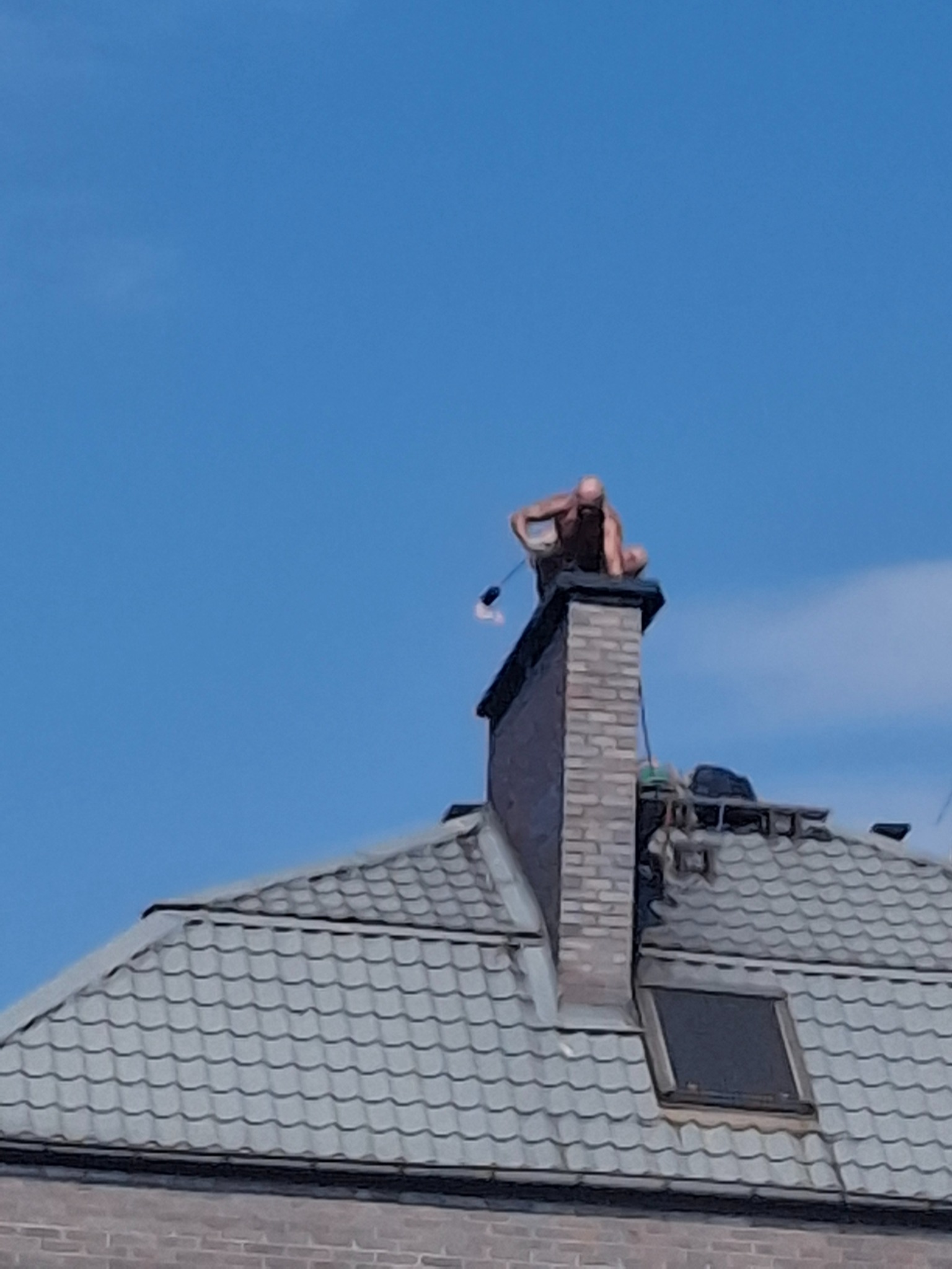 Mężczyzna siedzi na kominie z narzędziem na dachu pokrytym szarą dachówką, na tle błękitnego nieba. Nietypowe ujęcie z dołu.