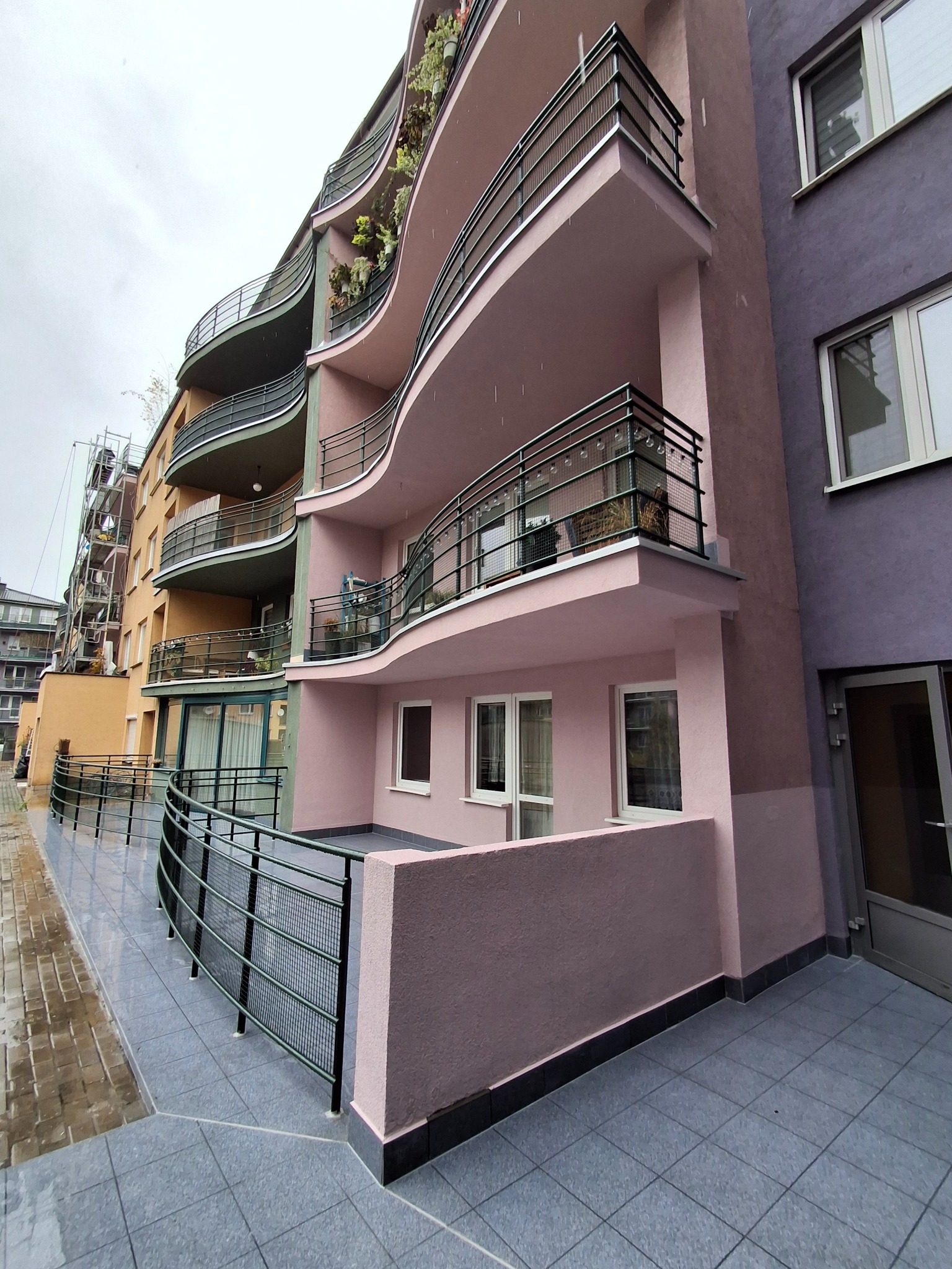 Wielopiętrowy budynek z balkonami o falistych liniach, pomalowany na odcienie różu i fioletu. Widoczne metalowe balustrady i rośliny na balkonach. Szara, deszczowa pogoda.