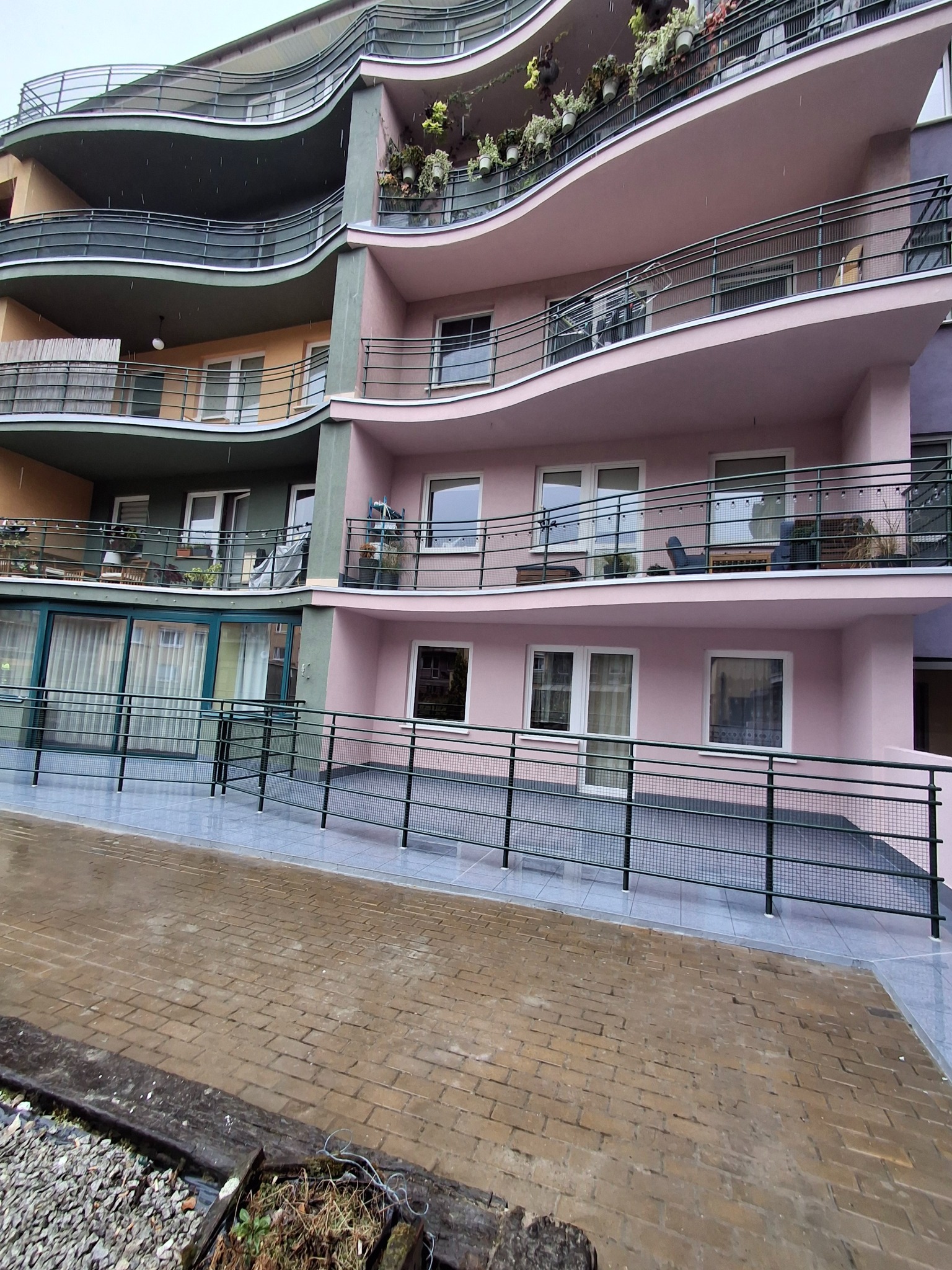 Wielopiętrowy budynek z falistymi balkonami w odcieniach różu i zieleni, widok z dołu na elewację i chodnik z kostki brukowej w deszczowy dzień.
