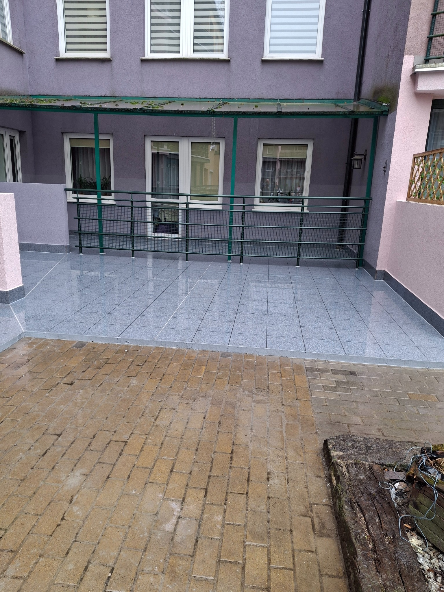 Balkon po remoncie z szarymi płytkami i zieloną balustradą. Widoczne okna budynku w tle. Na pierwszym planie kostka brukowa.