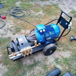 Olikamfram - Profesjonalna, mobilna myjka ciśnieniowa z silnikiem elektrycznym na trawie, z widocznymi złączkami i przewodem zasilającym. Obok wkrętarka akumulatorowa.