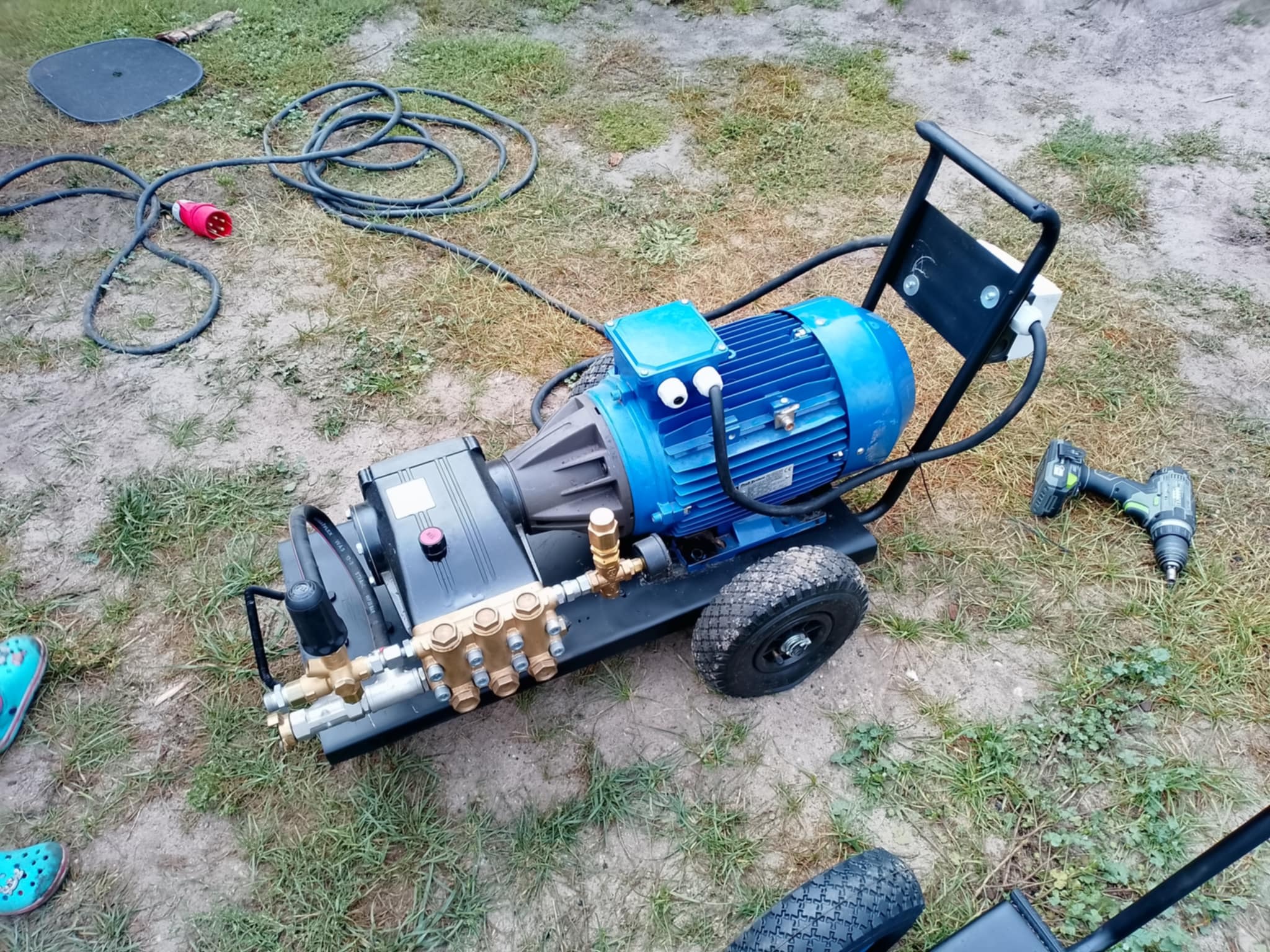 Profesjonalna, mobilna myjka ciśnieniowa z silnikiem elektrycznym na trawie, z widocznymi złączkami i przewodem zasilającym. Obok wkrętarka akumulatorowa.