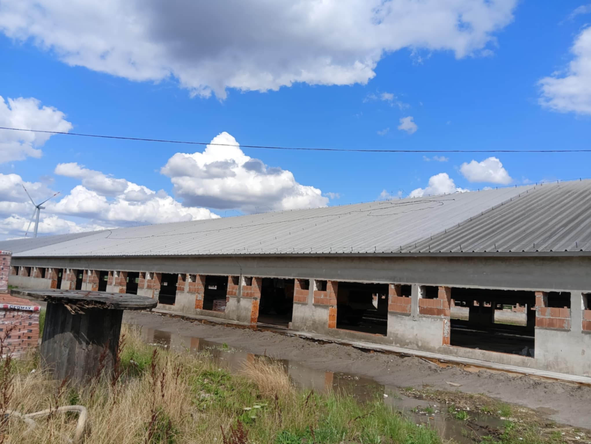 Budynek gospodarczy w trakcie budowy z widocznymi cegłami, szarym dachem i kałużami. W tle błękitne niebo z chmurami i turbina wiatrowa.