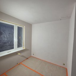 COMFORTbud KOSTIANTYN OVCHINNIKOV - Pokój po malowaniu ścian na szaro, okno zabezpieczone folią, podłoga zakryta kartonem i taśmą. Widoczny reflektor doświetlający.