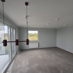 COMFORTbud KOSTIANTYN OVCHINNIKOV - Wnętrze w szarościach po malowaniu ścian, z widocznymi oknami, grzejnikiem i nowoczesną lampą wiszącą. Podłoga surowa, przygotowana do dalszych prac wykończeniowych.