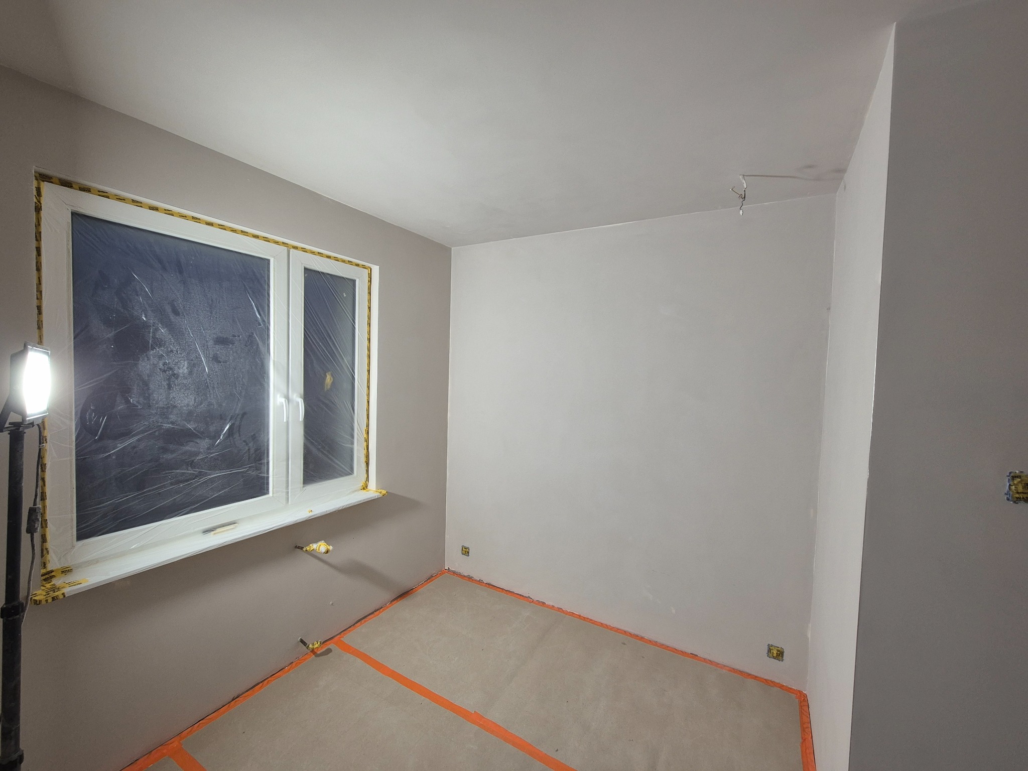 Pokój po malowaniu ścian na szaro, okno zabezpieczone folią, podłoga zakryta kartonem i taśmą. Widoczny reflektor doświetlający.