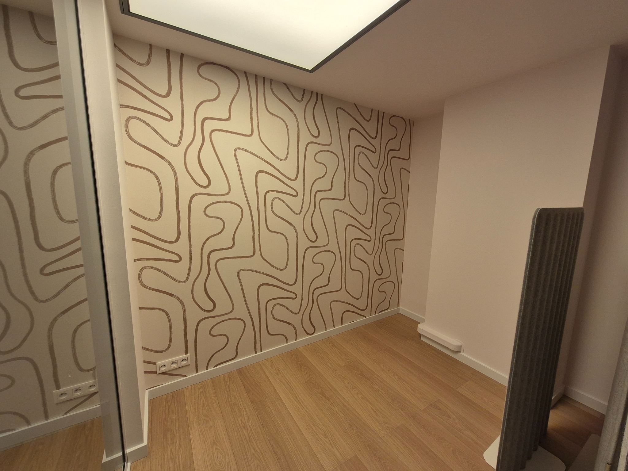 Pokój po remoncie: drewniana podłoga, ściana z wzorzystą tapetą w odcieniach beżu i brązu, nowoczesne oświetlenie sufitowe. Widoczne gniazdka elektryczne.