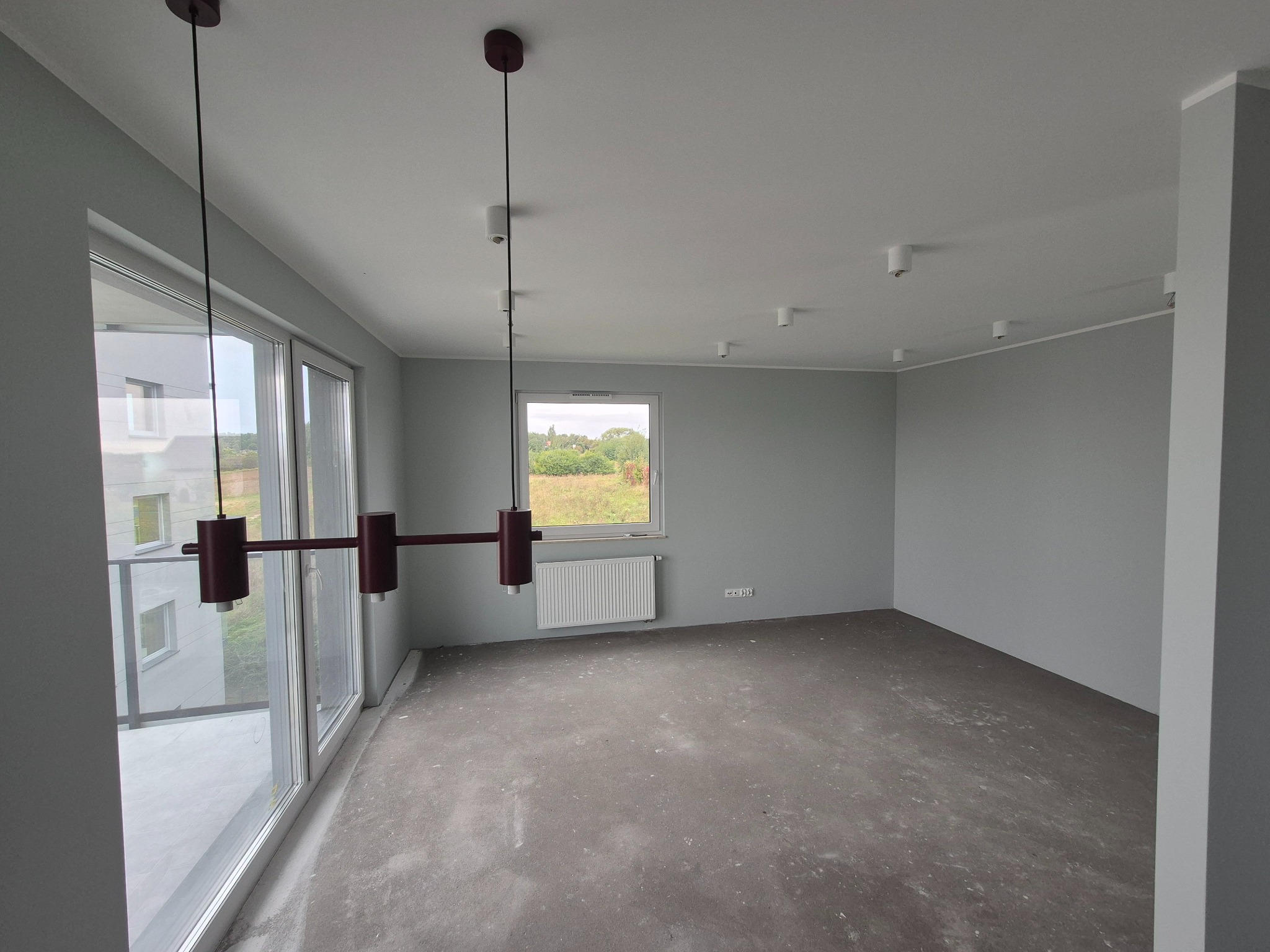 Wnętrze w szarościach po malowaniu ścian, z widocznymi oknami, grzejnikiem i nowoczesną lampą wiszącą. Podłoga surowa, przygotowana do dalszych prac wykończeniowych.