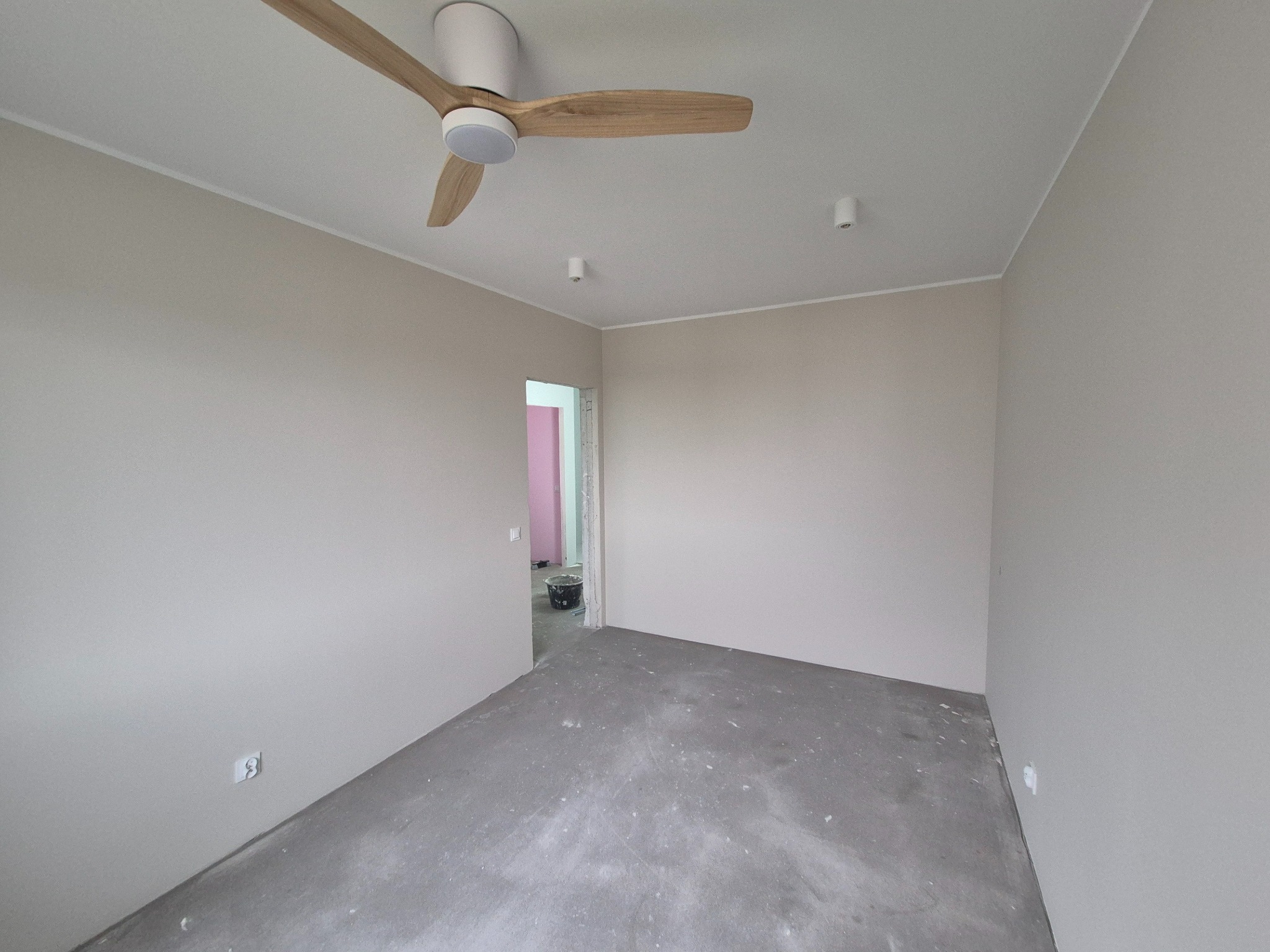 Pomalowane na beżowo wnętrze pokoju z betonową podłogą, widoczne gniazdko elektryczne, wentylator sufitowy i fragment różowej ściany w przejściu do innego pomieszczenia.