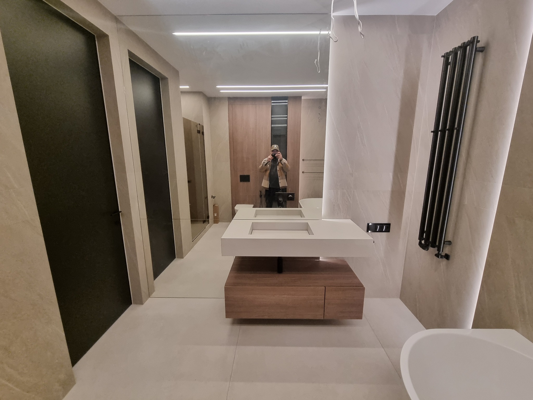 Nowoczesna łazienka z podwójną umywalką na szafce z drewna, czarnym grzejnikiem drabinkowym i lustrem odbijającym osobę robiącą zdjęcie. Minimalistyczny design.