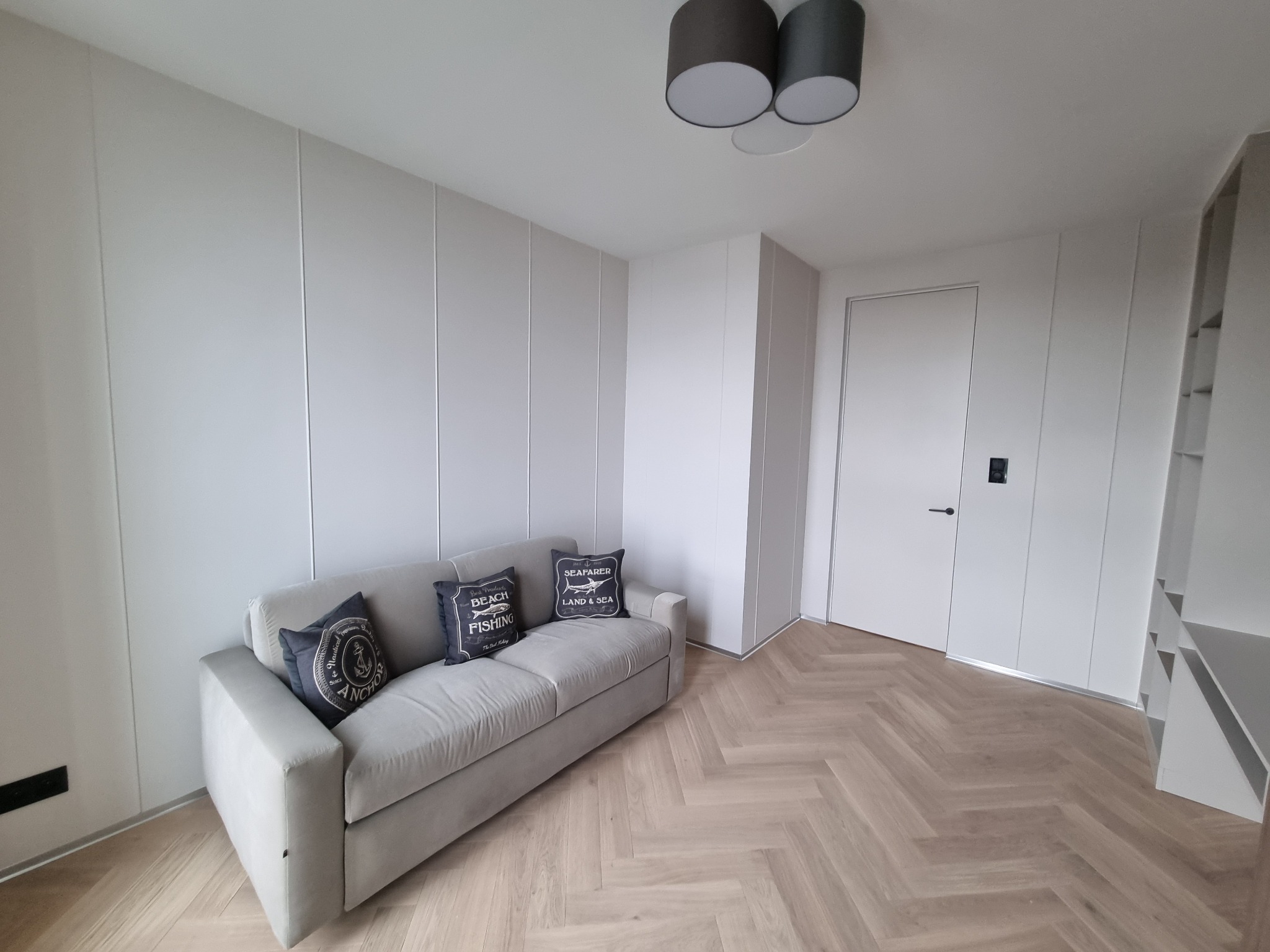 Nowoczesne wnętrze: szara sofa z dekoracyjnymi poduszkami na parkiecie w jodełkę, białe panele ścienne z subtelnymi pionowymi podziałami, minimalistyczne oświetlenie sufitowe.