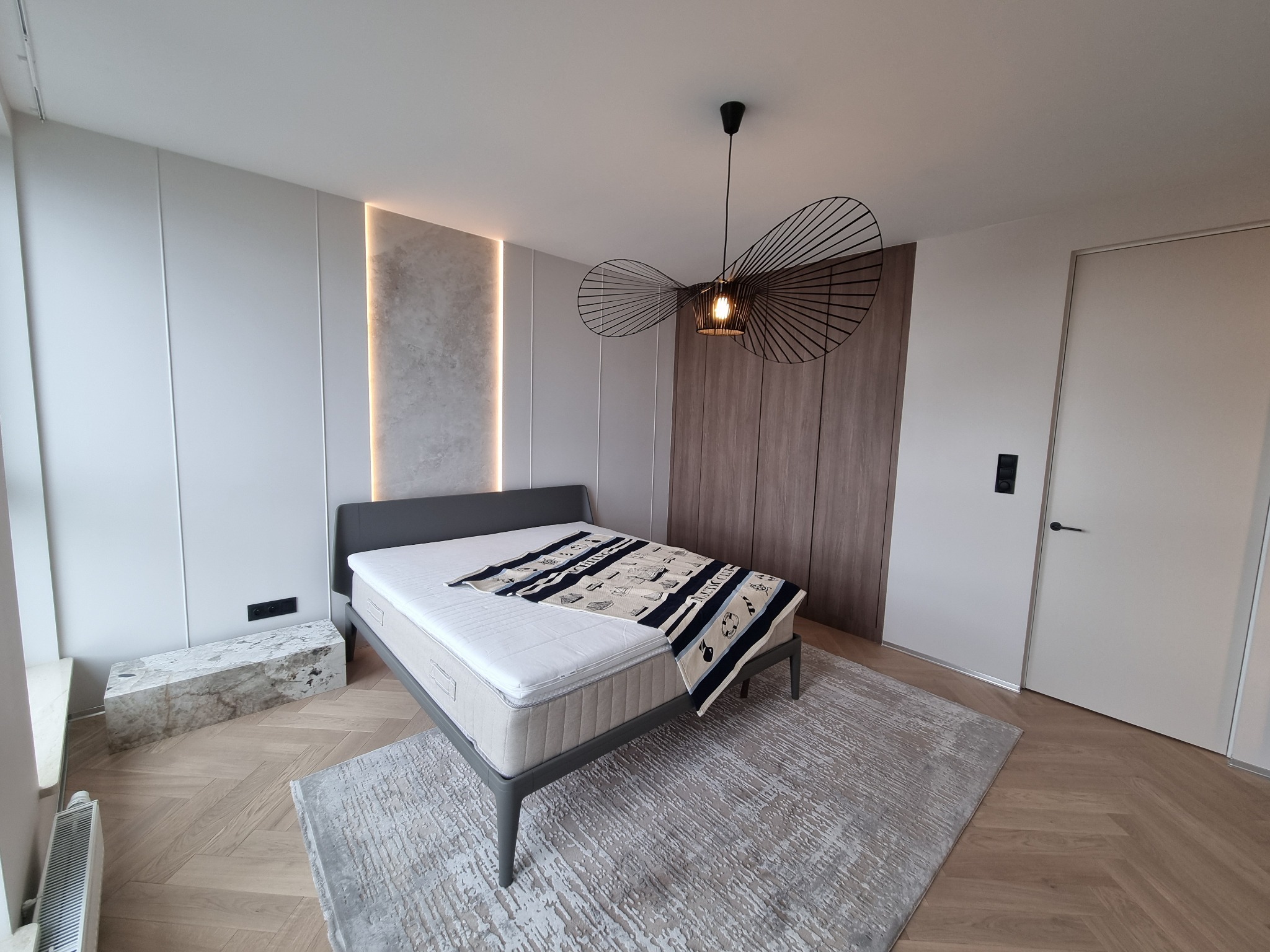 Nowoczesna sypialnia z łóżkiem, designerską lampą i drewnianą podłogą w jodełkę. Minimalistyczny wystrój z akcentami marmuru i subtelnym oświetleniem.