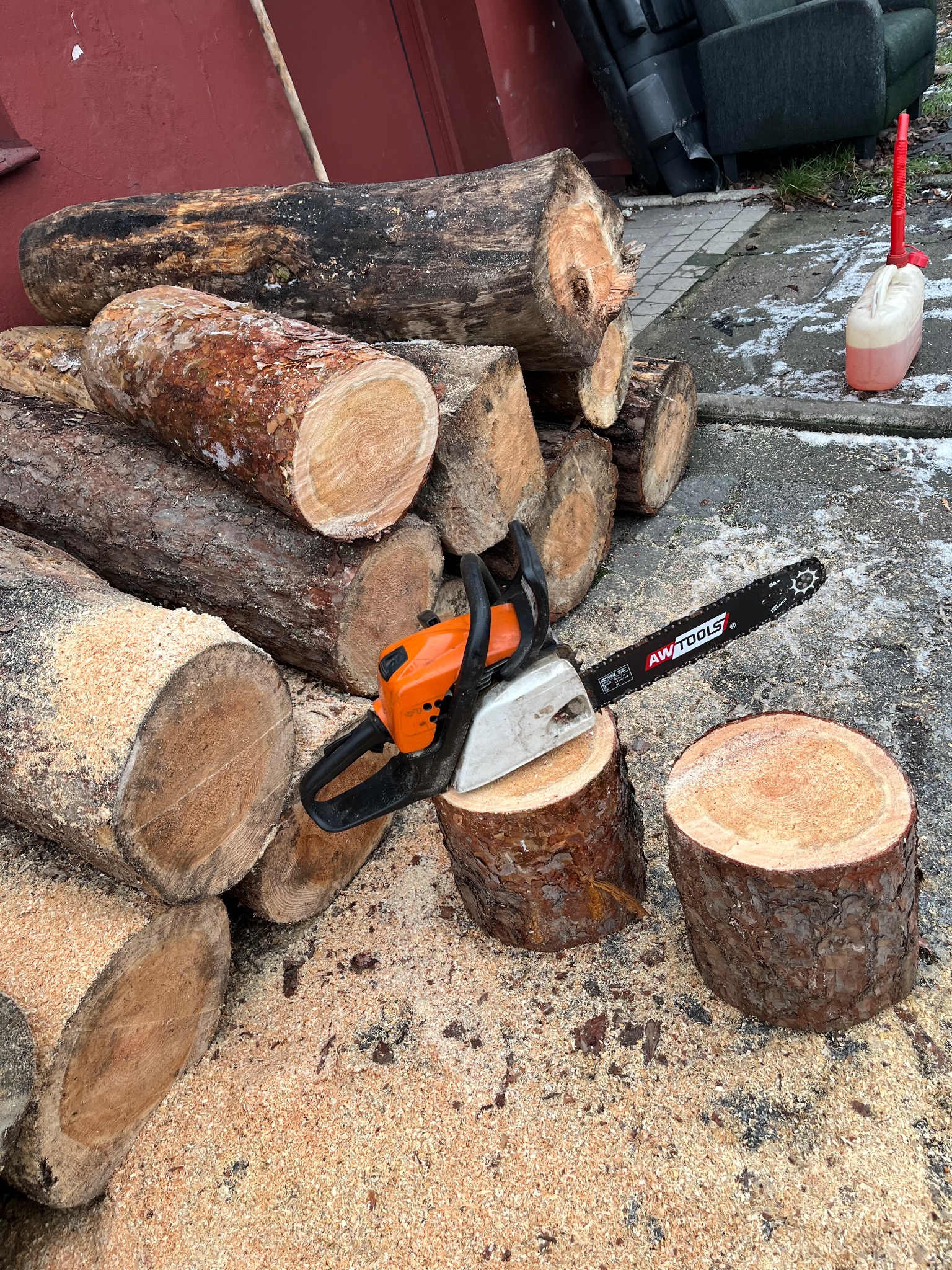 Stos pociętych kloców drewna z piłą łańcuchową i kanistrem na tle betonowej nawierzchni i czerwonej ściany. Widoczne świeże trociny.